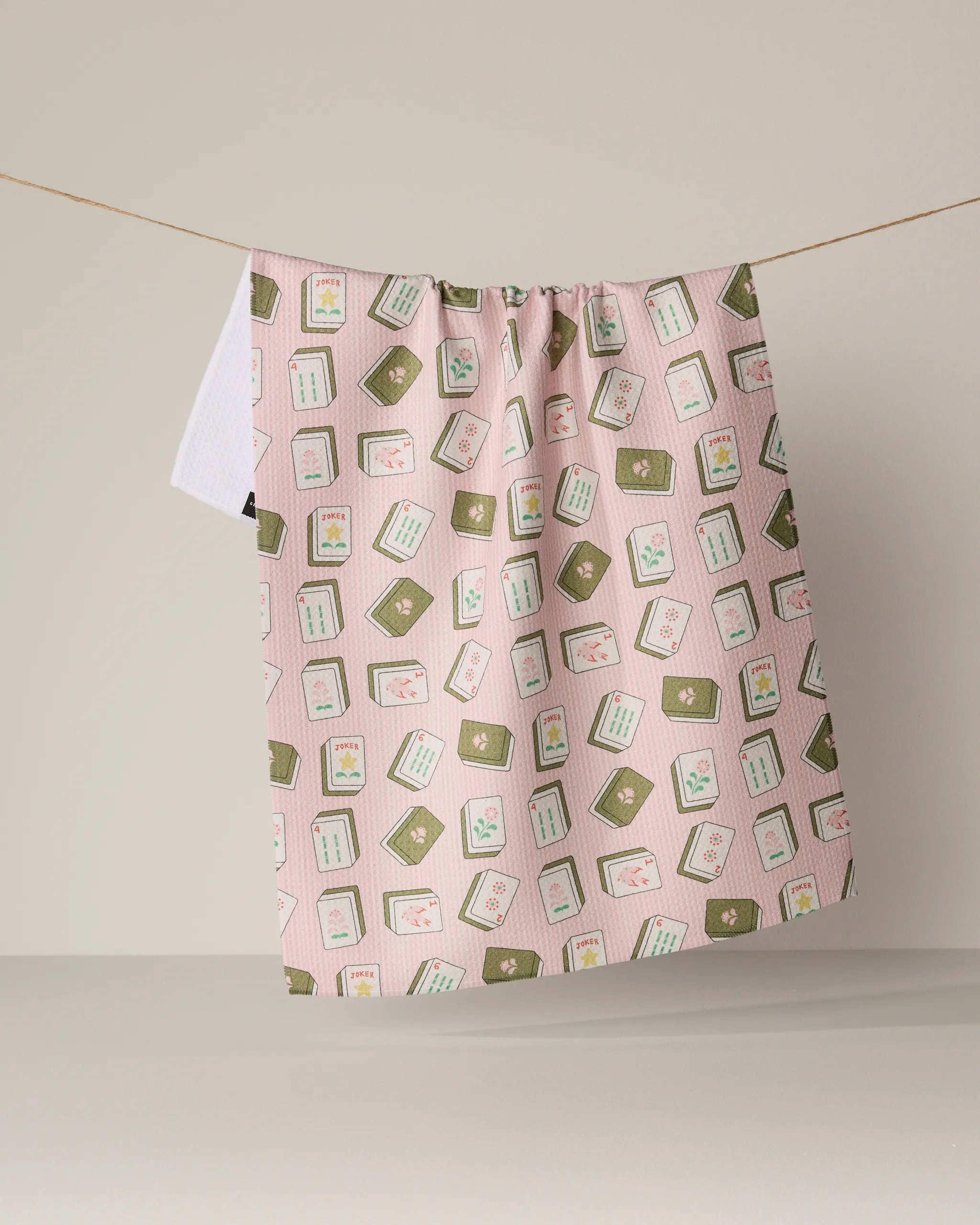 A "Flower Tiles" kitchen towel by Geometry, featuring an eco-friendly design in pink with green and white square patterns and small floral accents, hangs on a clothesline against a plain beige background.