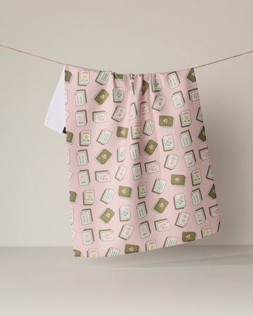 A "Flower Tiles" kitchen towel by Geometry, featuring an eco-friendly design in pink with green and white square patterns and small floral accents, hangs on a clothesline against a plain beige background.