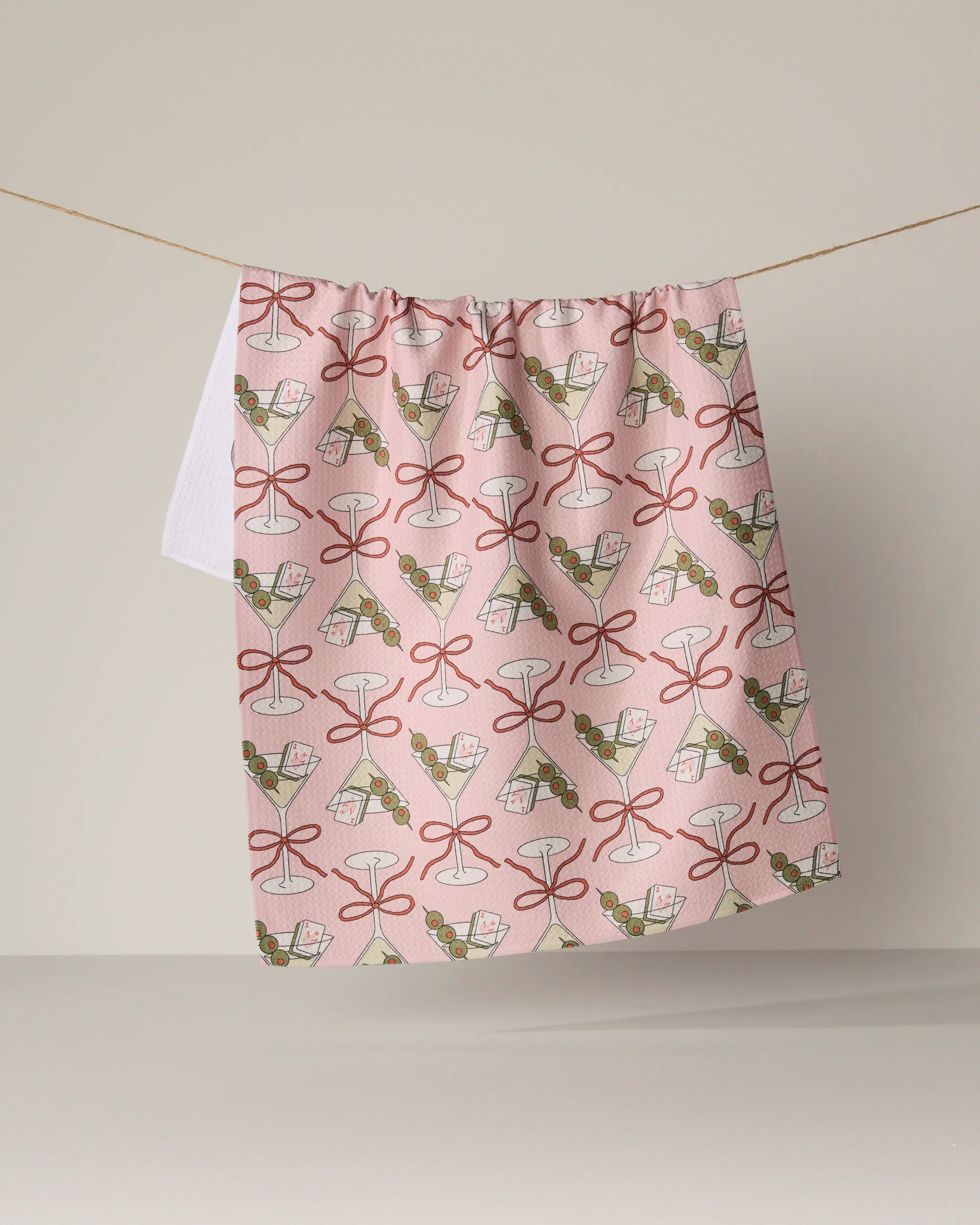 The Geometry Flower-tinis tea towel, featuring a pink background and a repeating pattern of cocktail glasses, olives, and bows, hangs on a string against a neutral backdrop. This absorbent dish towel brings playful charm to your kitchen while being designed for everyday use.