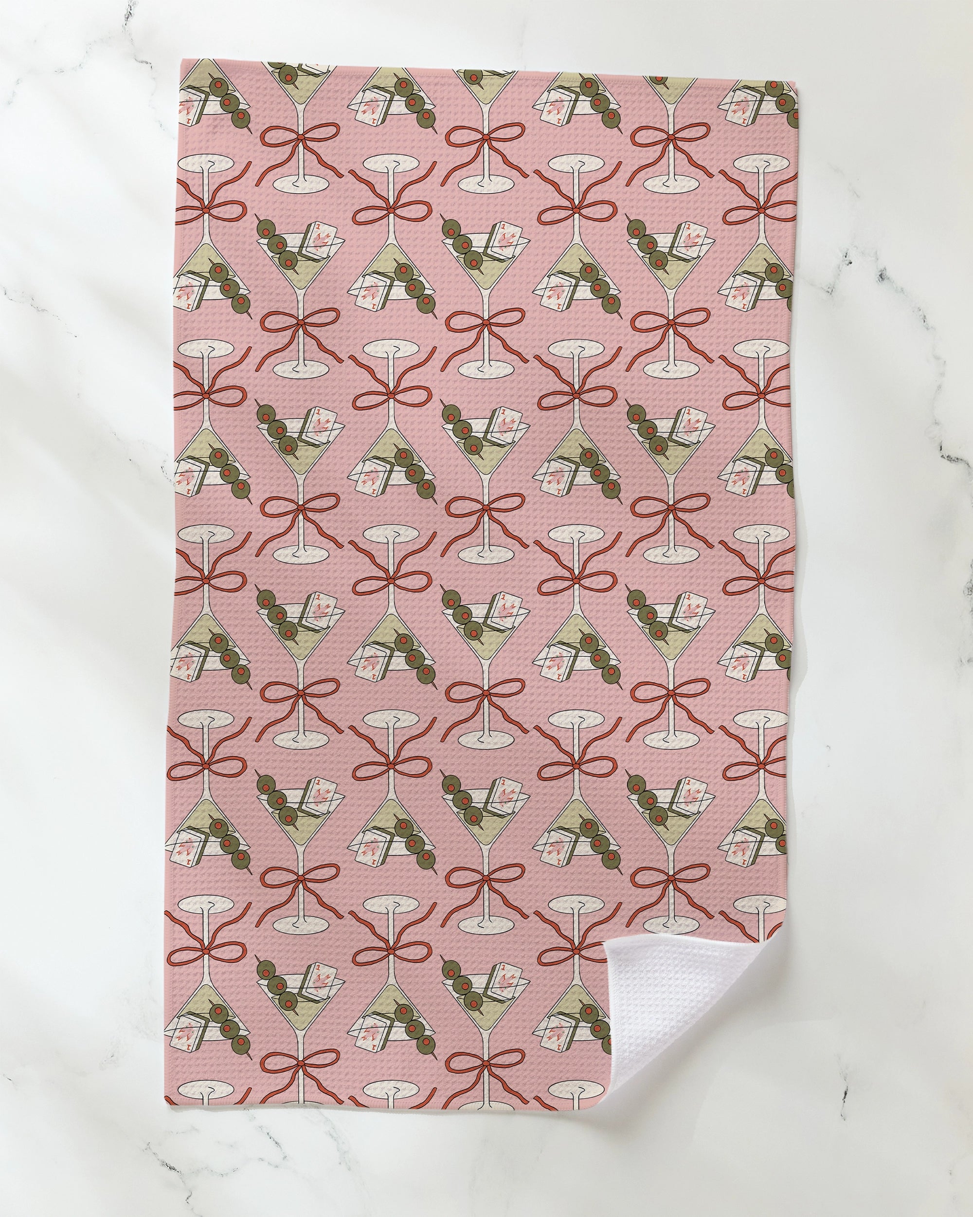 A pink "Flower-tinis" kitchen tea towel by Geometry, featuring a repeating pattern of martini glasses, olives, and cocktail napkins, crafted from recycled materials and shown on a white marble surface with one corner slightly folded.