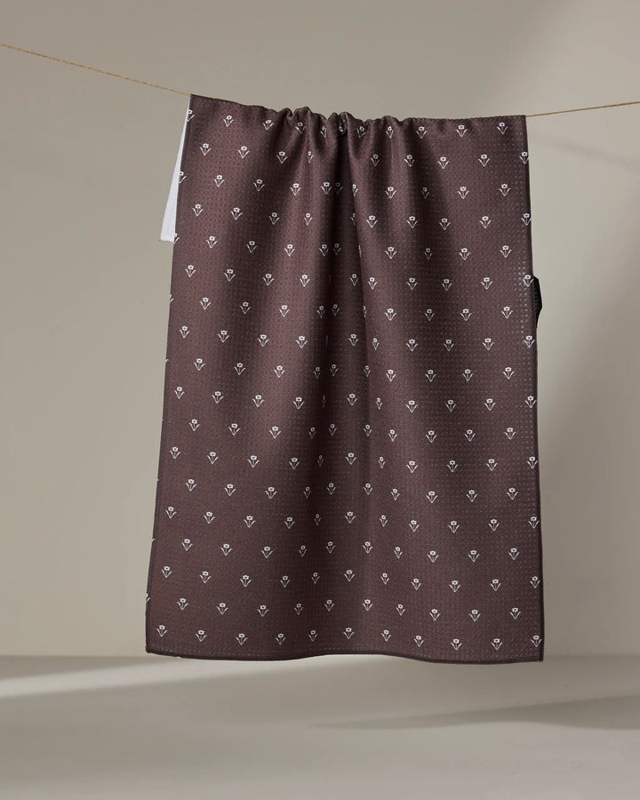 The Buy Guide’s Flowerfield Chocolate tea towel, crafted from recycled materials and featuring white diamond and floral patterns, hangs over a clothesline against a neutral background.