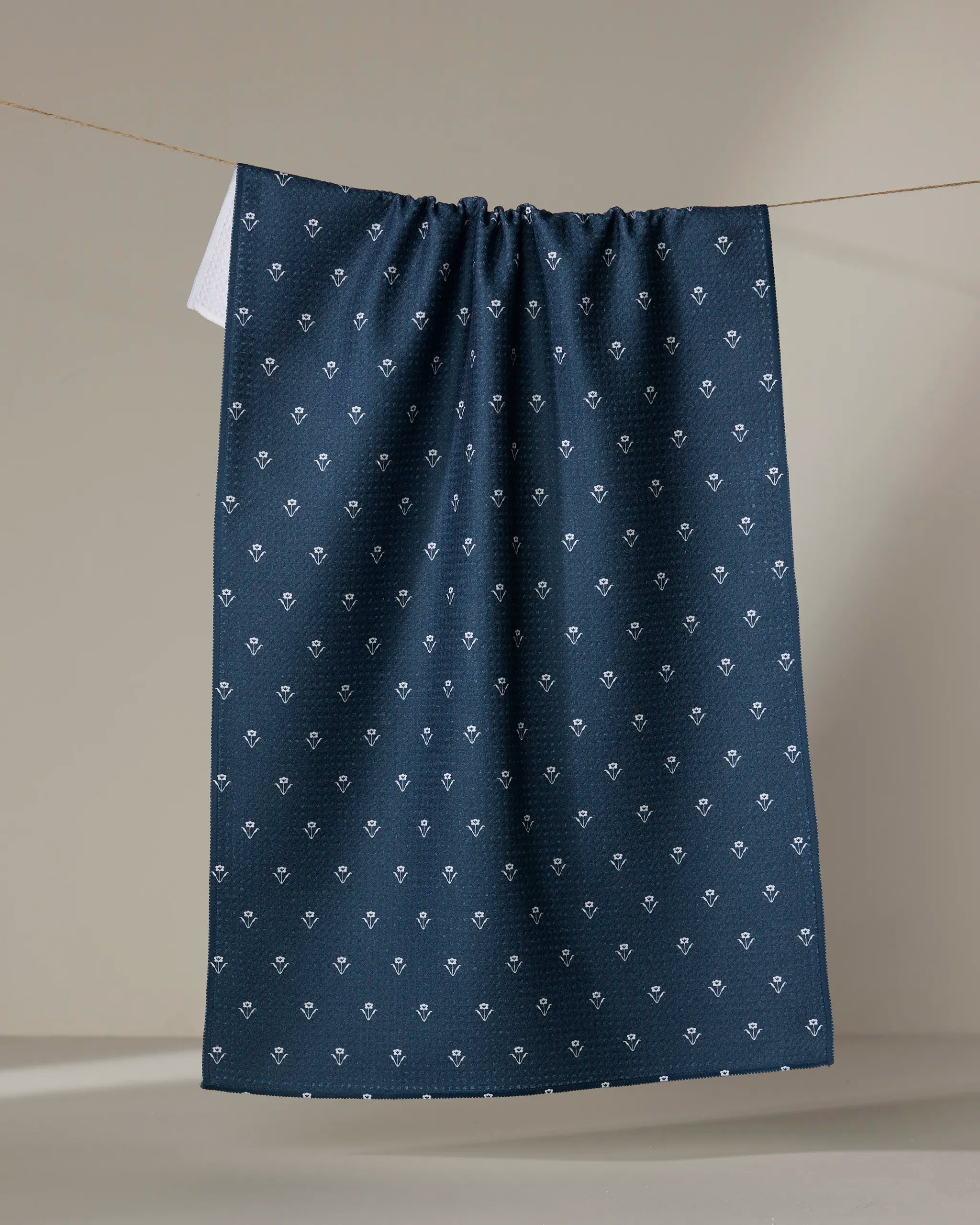 The Buy Guide’s Flowerfield Navy tea towel, featuring a dark blue pattern with small white geometric shapes, hangs on a string against a neutral background.