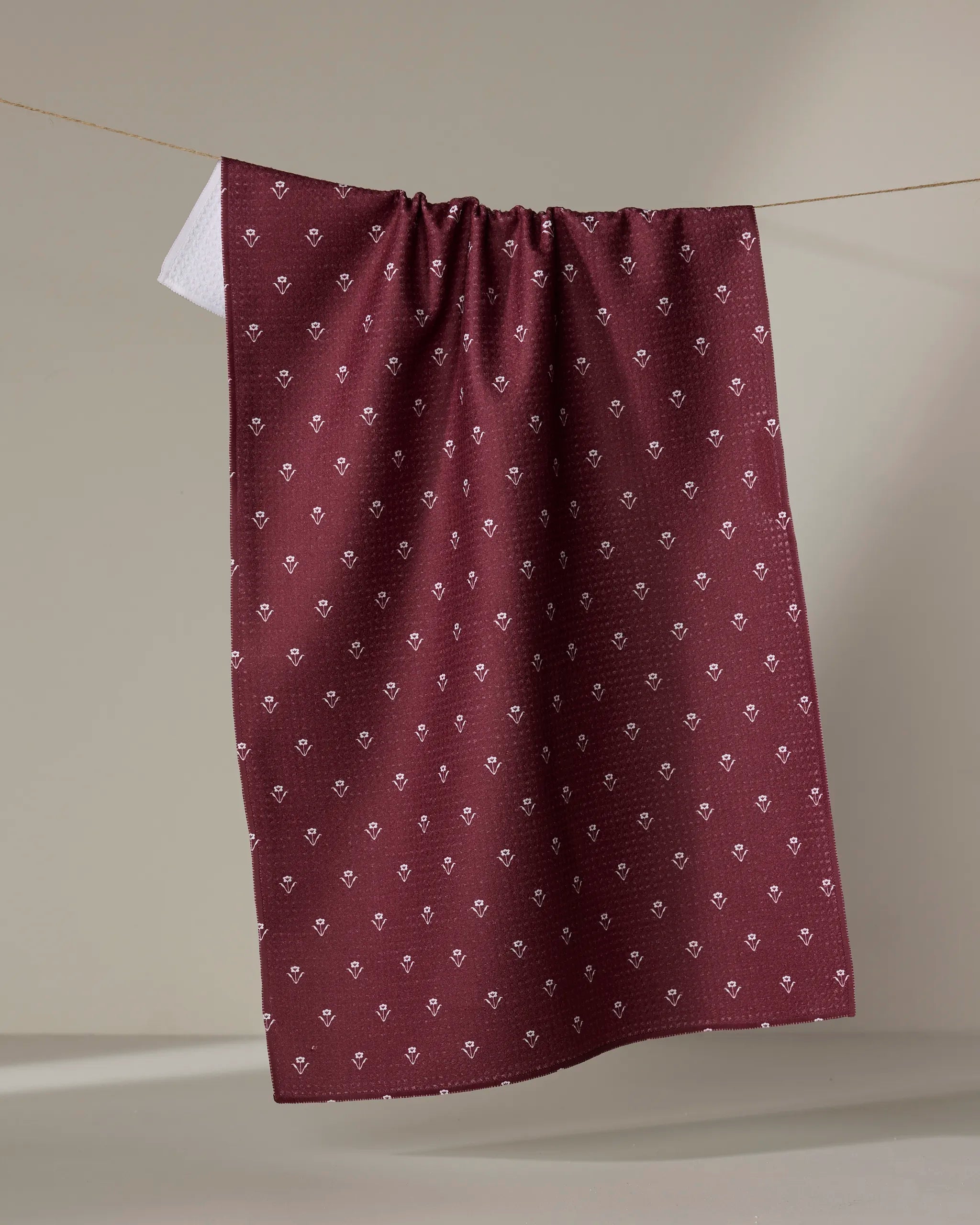 The Buy Guide’s Flowerfield Wine tea towel features a maroon color with a small white floral pattern, hanging on a string against a neutral background—perfect for adding charm to your kitchen decor.