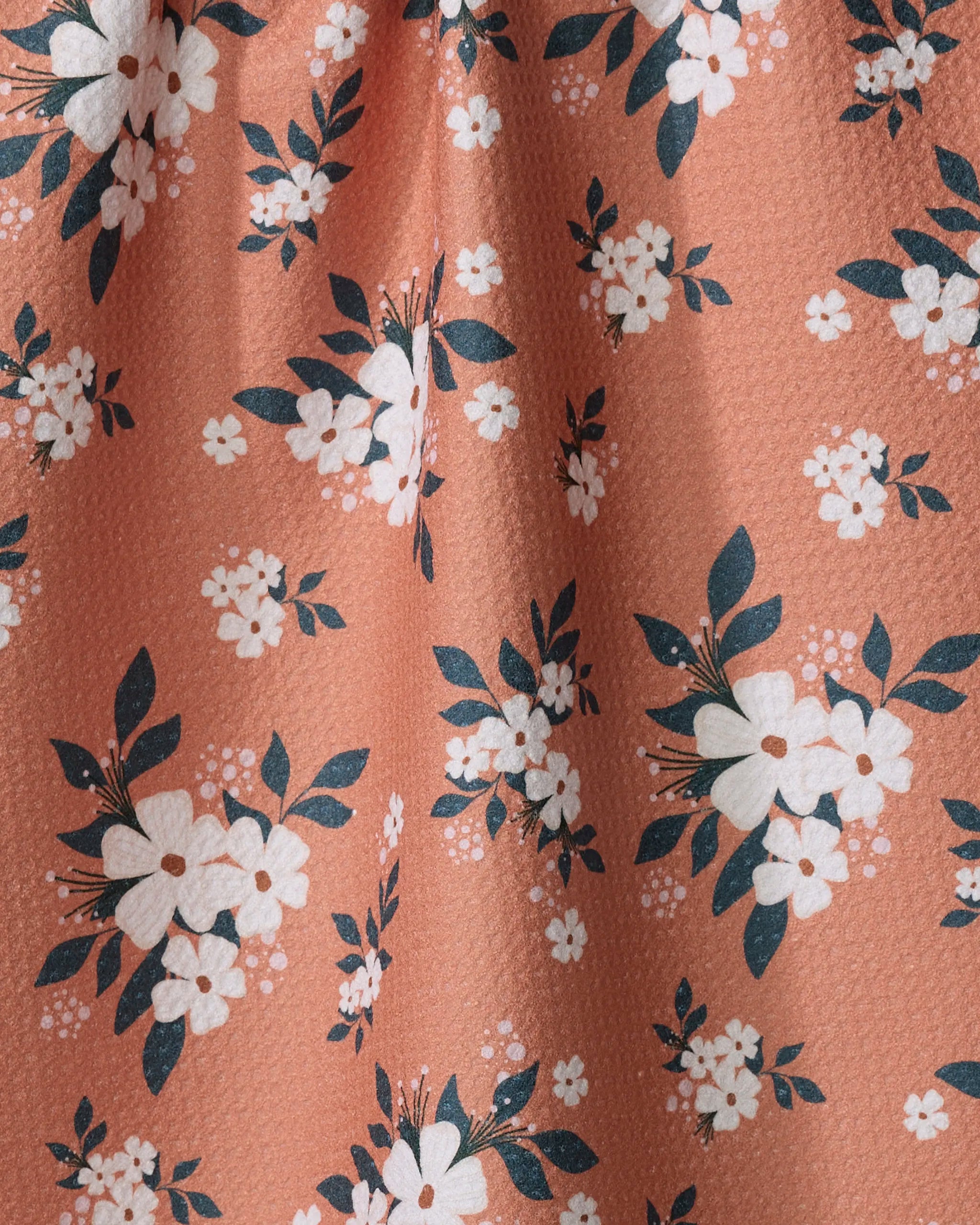 A close-up of the "Flowers in the Field" tea towel by Charlie Rowan Designs shows peach-colored, textured fabric with white floral patterns and dark green leaves, softly gathered to form gentle folds and shadows.
