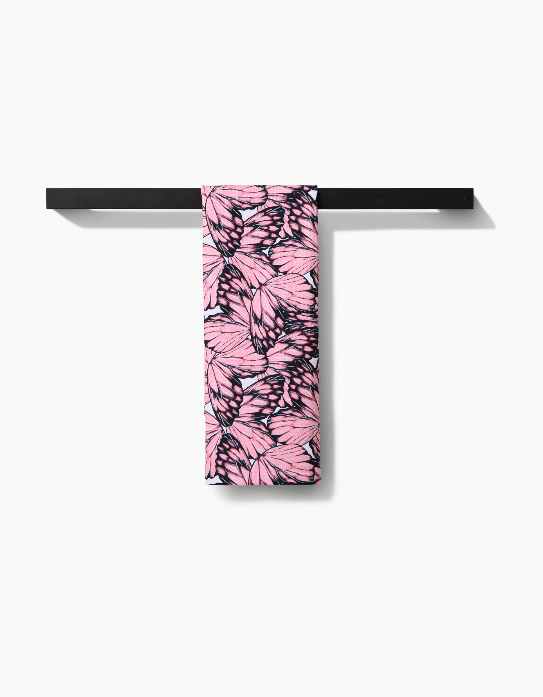 The Flutter by Butterfly tea towel by Aubrey Fairchild, featuring a black leaf pattern on pink recycled fabric, is displayed draped over a minimalist black towel rack against a white background.