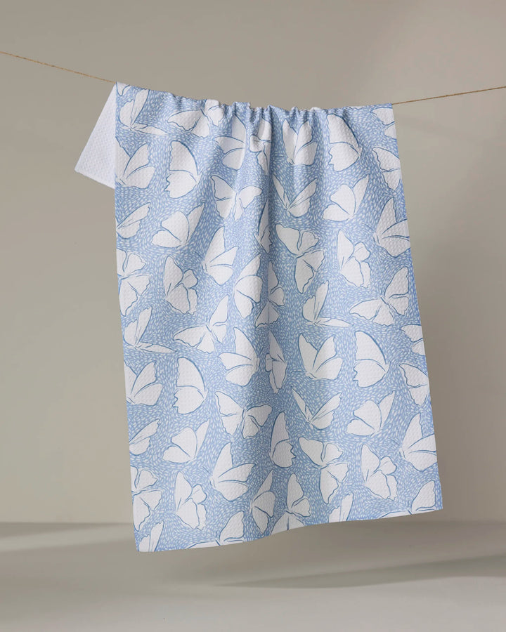 The Fly Away Butterfly tea towel by Hufton Studio features a white butterfly design on light blue fabric, hanging from a string against a neutral background. It gathers at the top and drapes down, highlighting its quick-drying quality.
