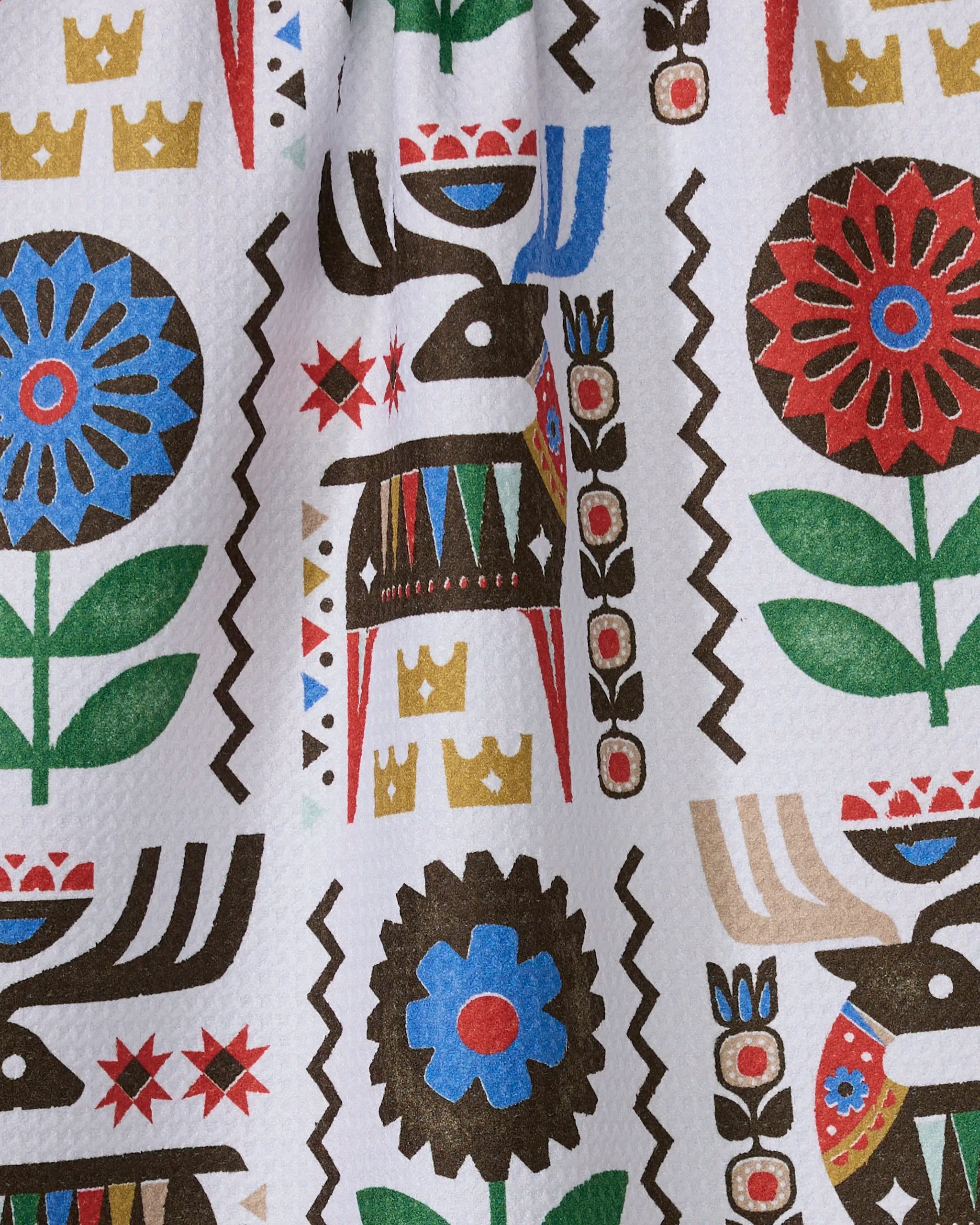 The Geometry "Folk Deer" tea towel features a colorful, folk art-style pattern with stylized animals, flowers, and crowns on textured white fabric made from ultra-absorbent recycled materials.