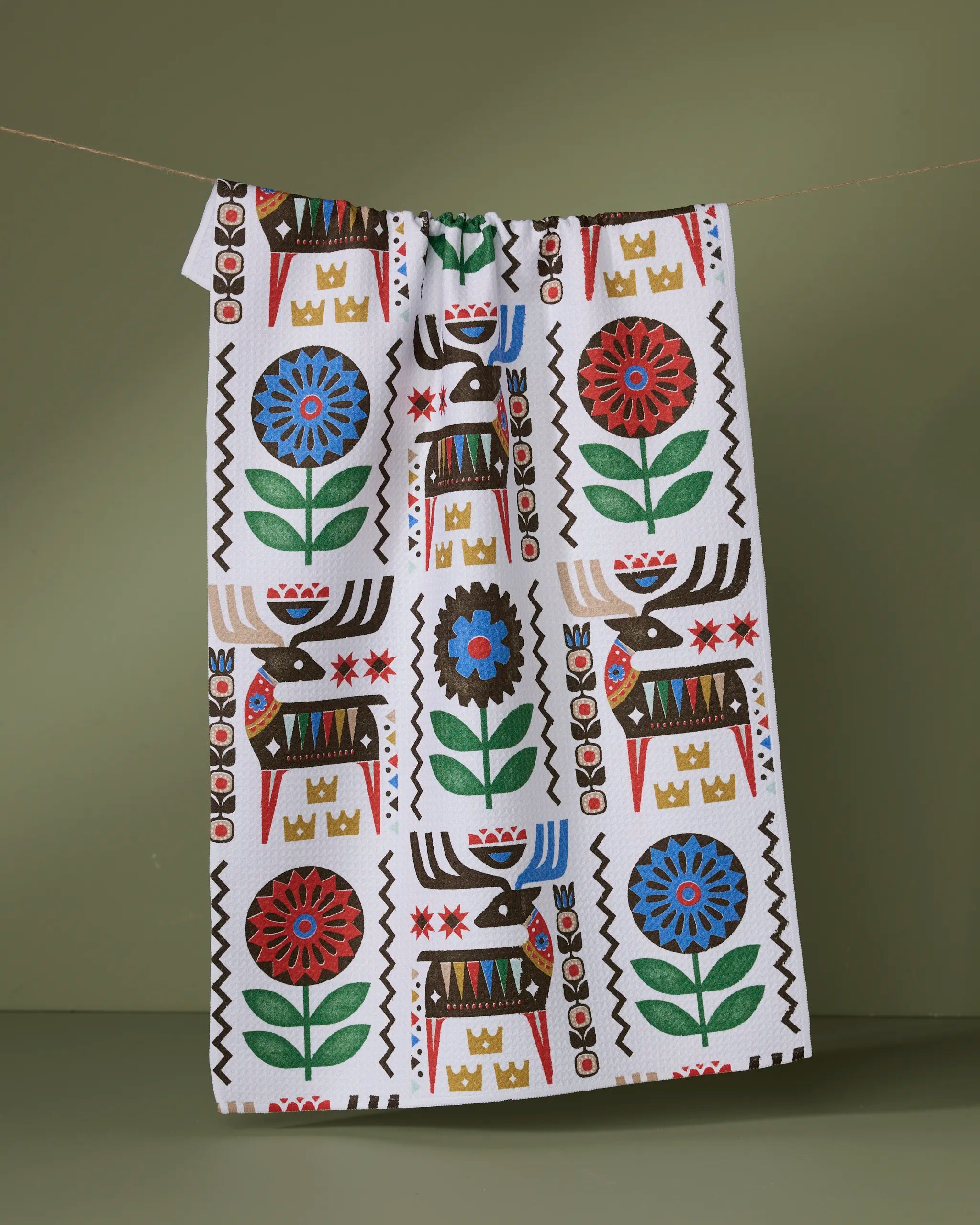 The Geometry Folk Deer tea towel, made from recycled materials, displays a vibrant folk-inspired pattern with brown reindeer, red and blue flowers, green leaves, and gold crowns against a muted green backdrop.