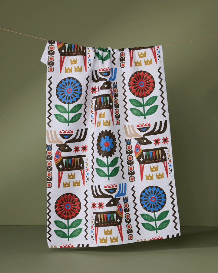 The Geometry Folk Deer tea towel, made from recycled materials, displays a vibrant folk-inspired pattern with brown reindeer, red and blue flowers, green leaves, and gold crowns against a muted green backdrop.