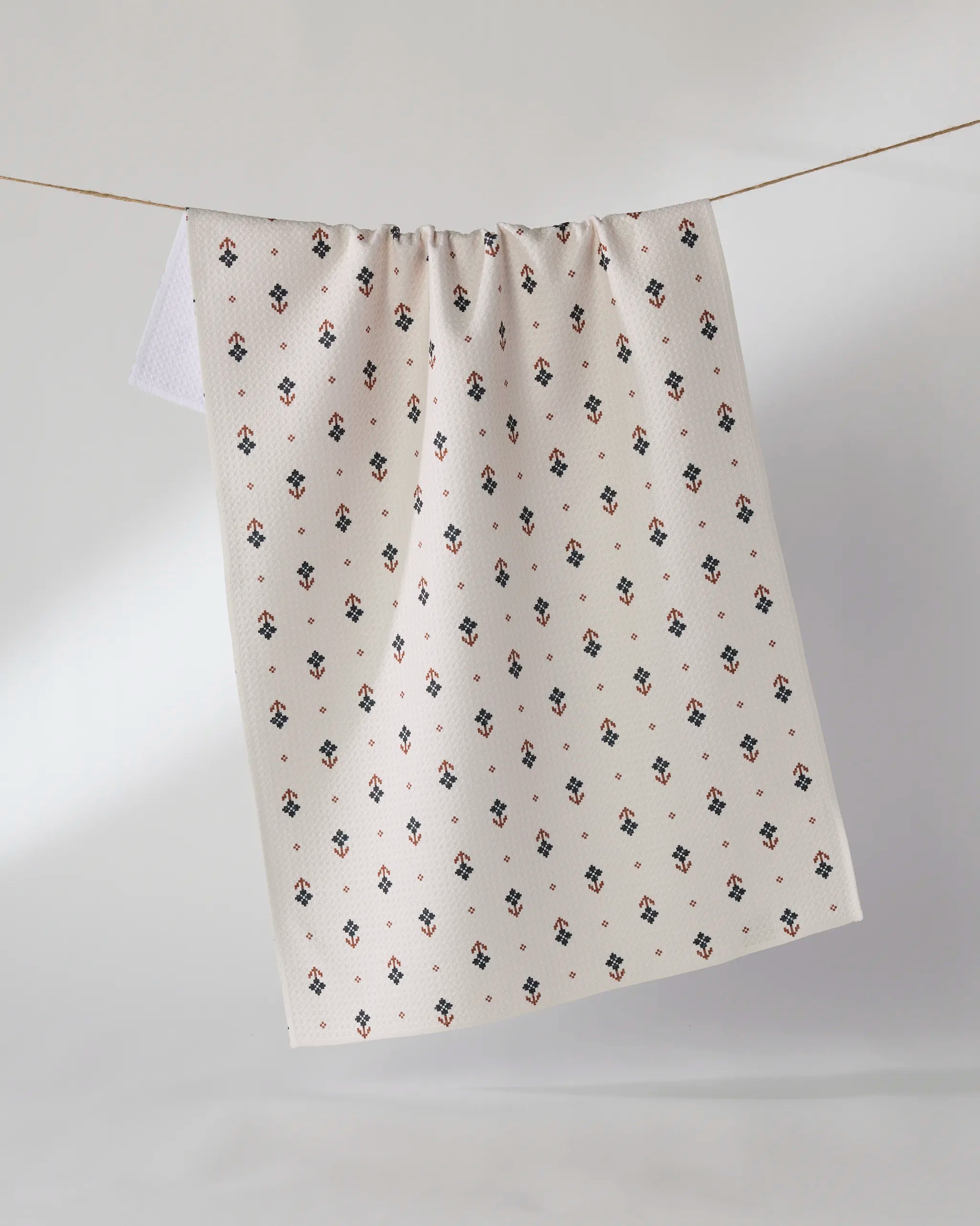 The Geometry Folk Pixel White kitchen towel, featuring an eco-friendly design with a small black and red floral pattern, hangs on a thin string against a plain light background.