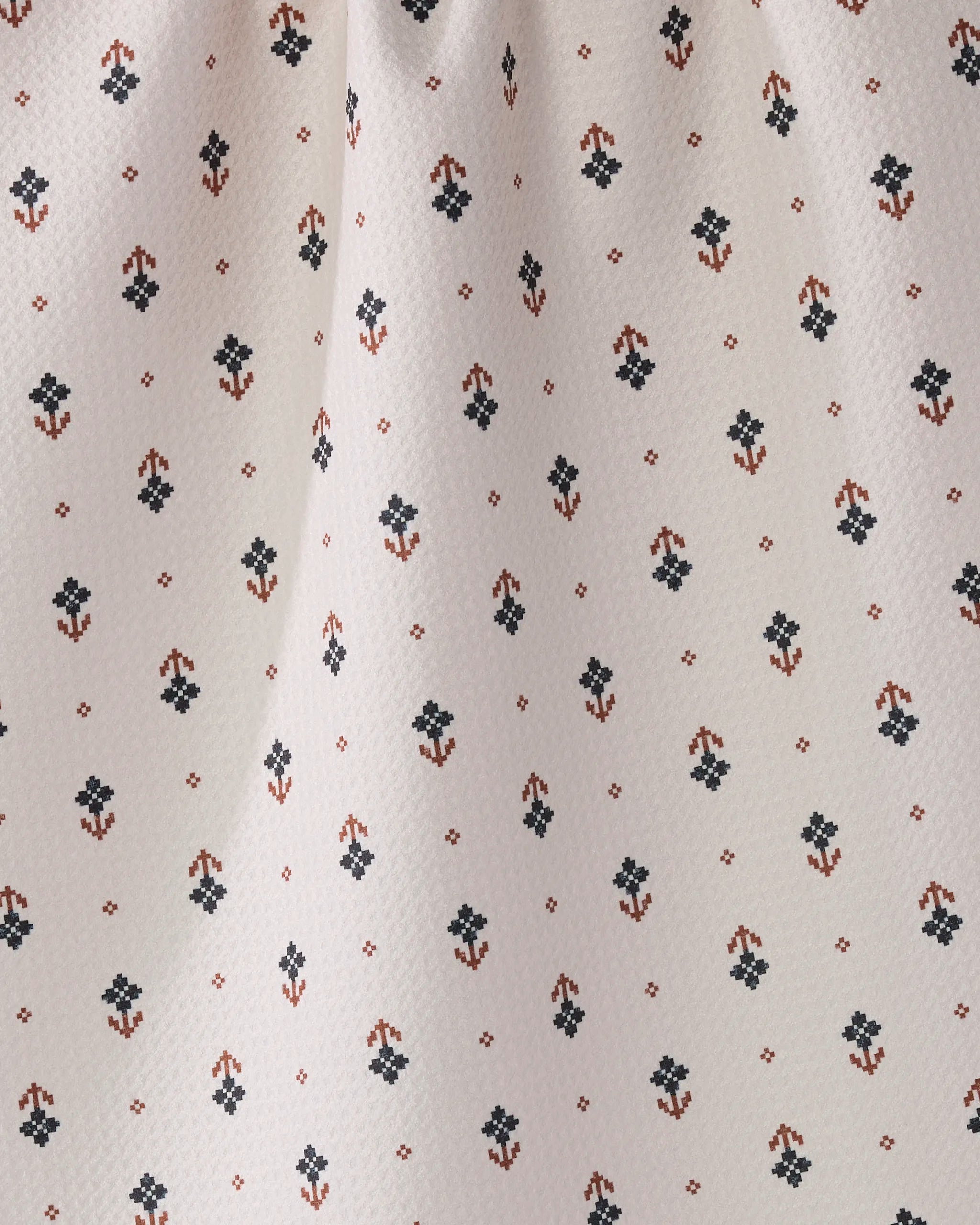 A close-up of Geometry’s Folk Pixel White, an eco-friendly textured fabric with small black and rust red floral patterns arranged in a grid—ideal as a tea towel, showing gentle folds and soft shadows.