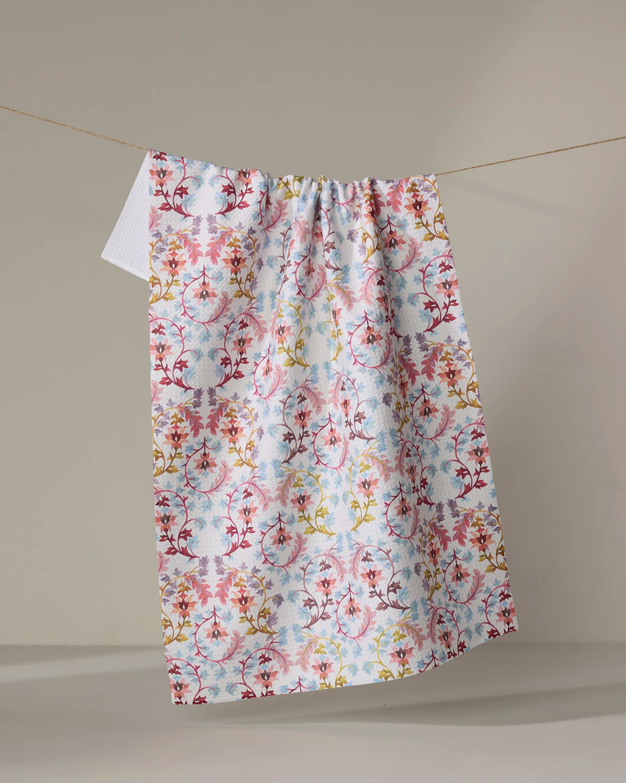 The “Folk Tapestry” kitchen towel by Maayan Alper-Swan, featuring eco-friendly fabric and vibrant red, yellow, and blue floral patterns, hangs on a clothesline against a plain beige background.