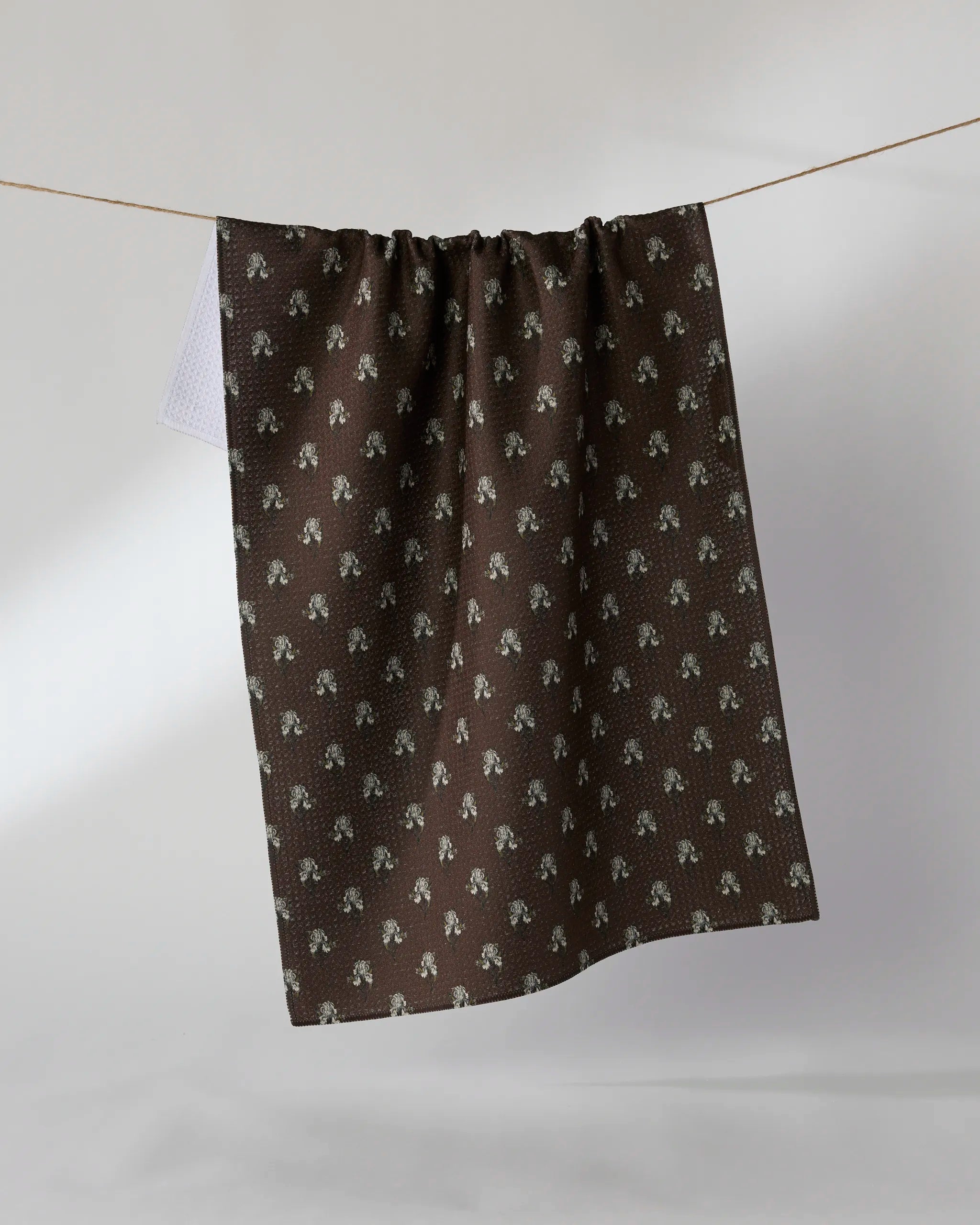 The Geometry Foraged recycled kitchen towel, featuring a small repeating floral pattern, hangs on a clothesline against a plain light gray background.
