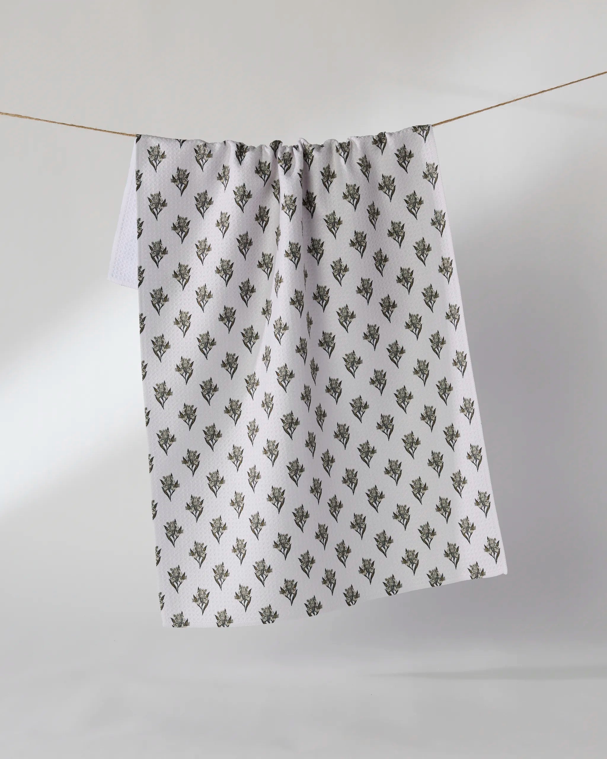 The Geometry Foraged towel, ultra-absorbent and white with a delicate floral pattern, hangs on a slim clothesline against a plain light background.