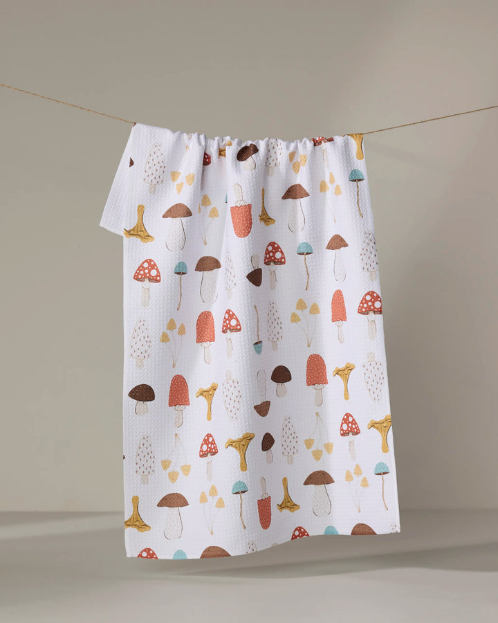 The "Foraging" recycled kitchen towel by Sara Mari, decorated with red, brown, yellow, and blue mushrooms, hangs on a clothesline against a neutral background.