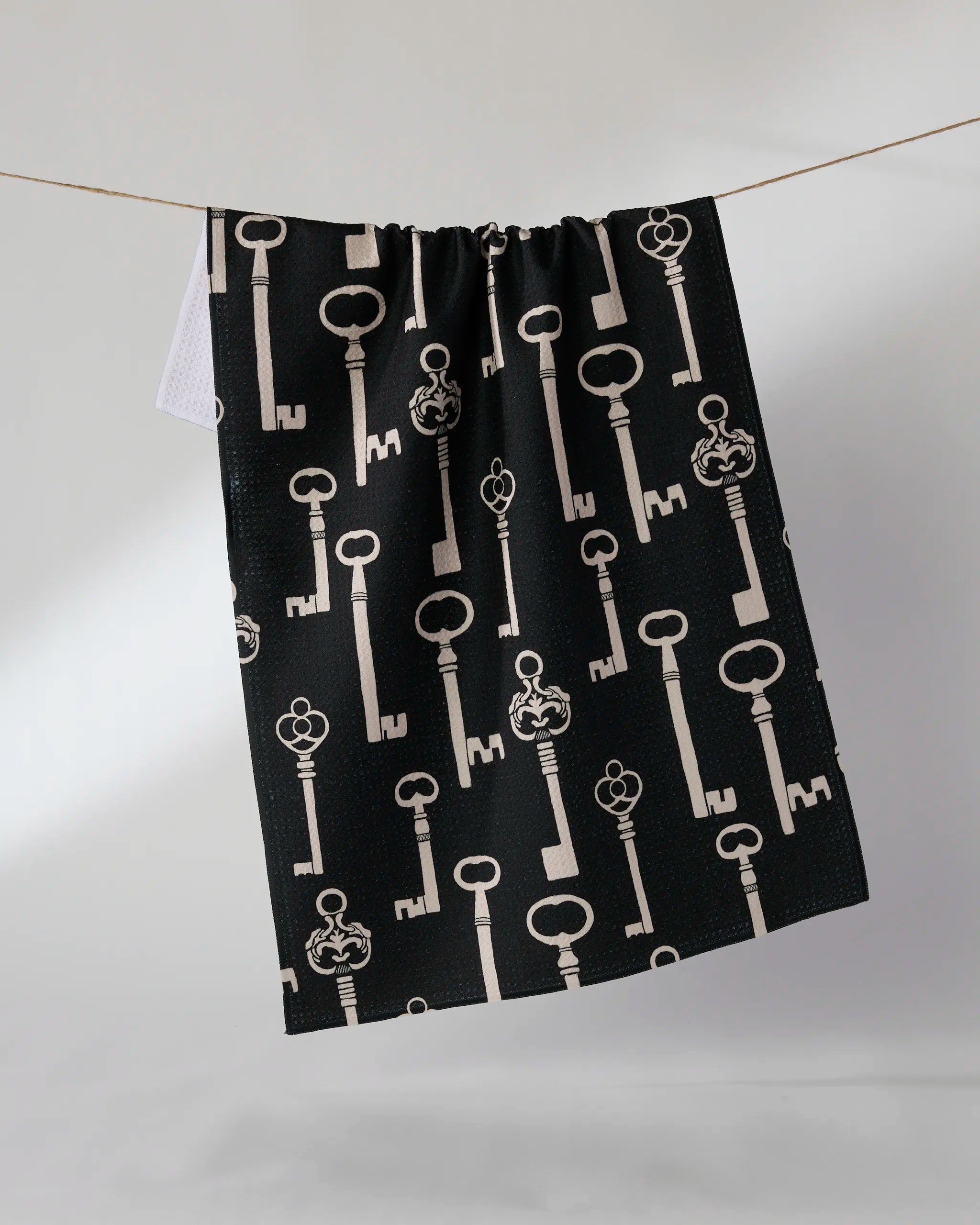 The "Forbidden Doors" tea towel by Geometry, in black with a vintage-style beige key pattern, hangs on a line against a plain, light background.