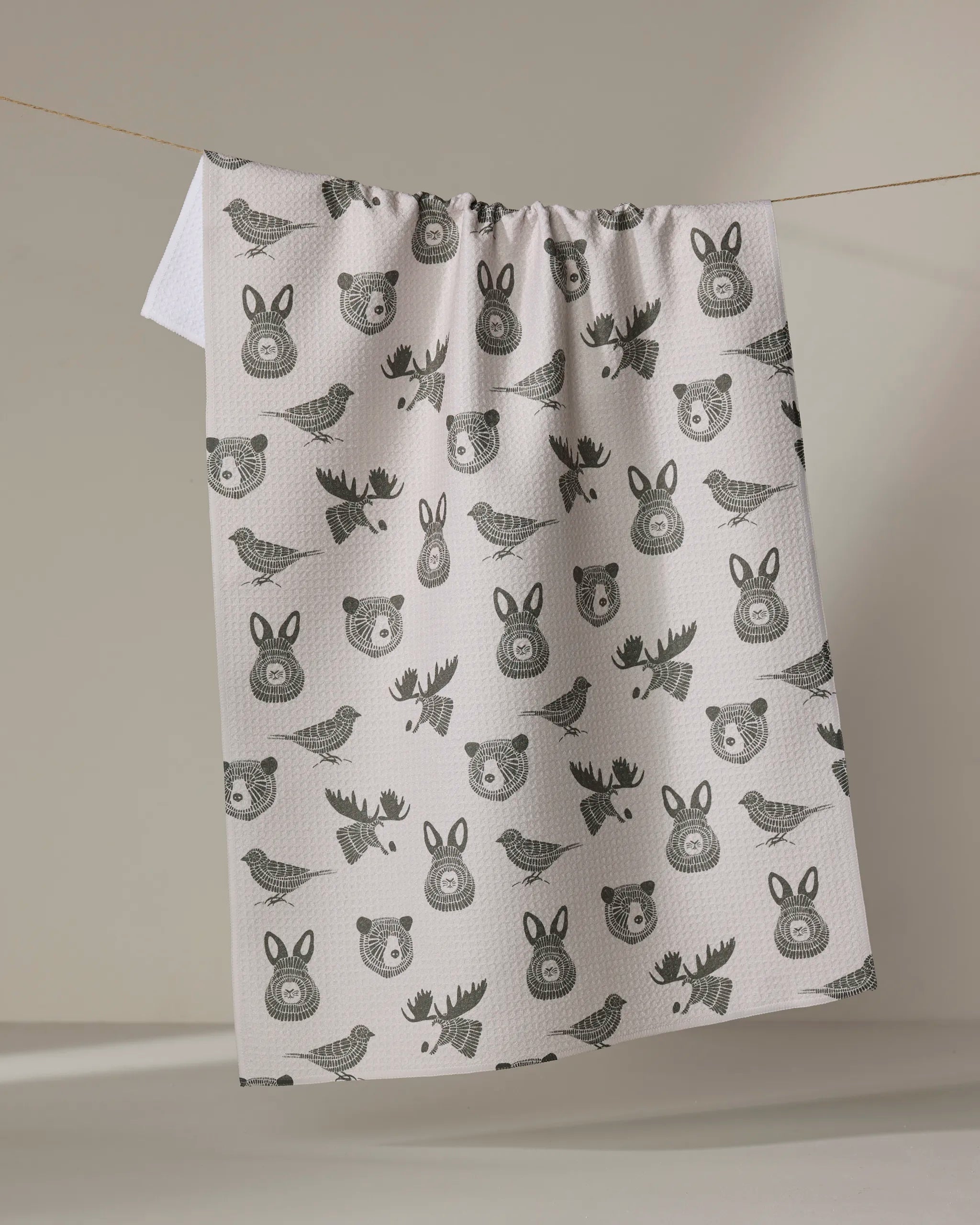 The "Forest Animals" tea towel by Rosa Kichline, featuring black prints of bears, birds, moose, and rabbit faces on white fabric, hangs on a clothesline against a softly lit plain background.