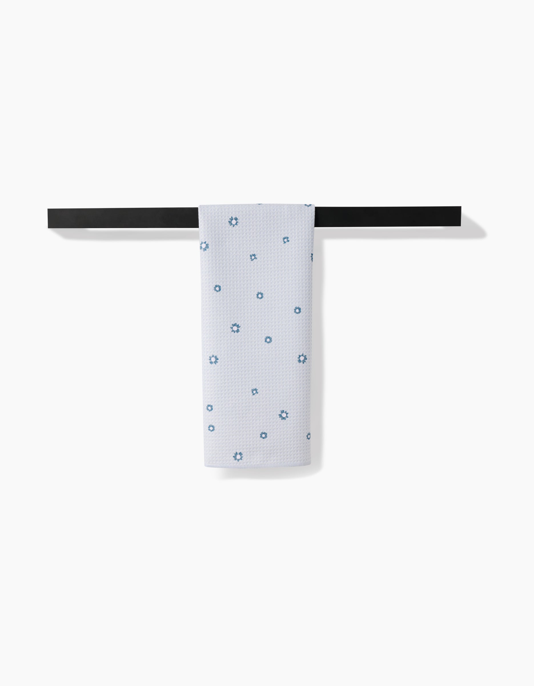 The Forest Daisy tea towel by Coit Creative, made from recycled materials and featuring small blue flowers, is draped over a black bar with a plain white background.