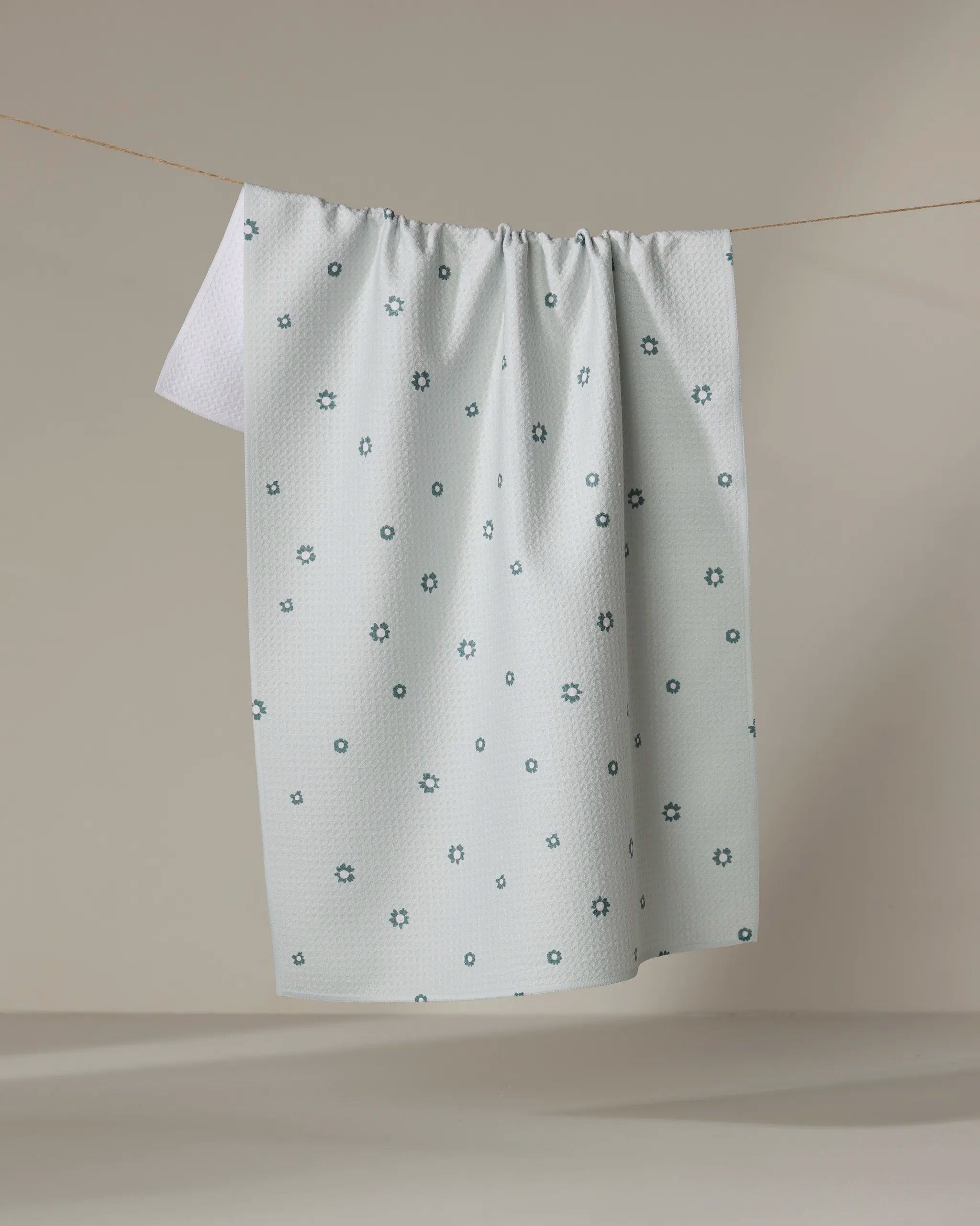 The Forest Daisy towel by Coit Creative, featuring a white ultra-absorbent textured surface and small blue geometric floral patterns, hangs on a clothesline against a neutral backdrop.