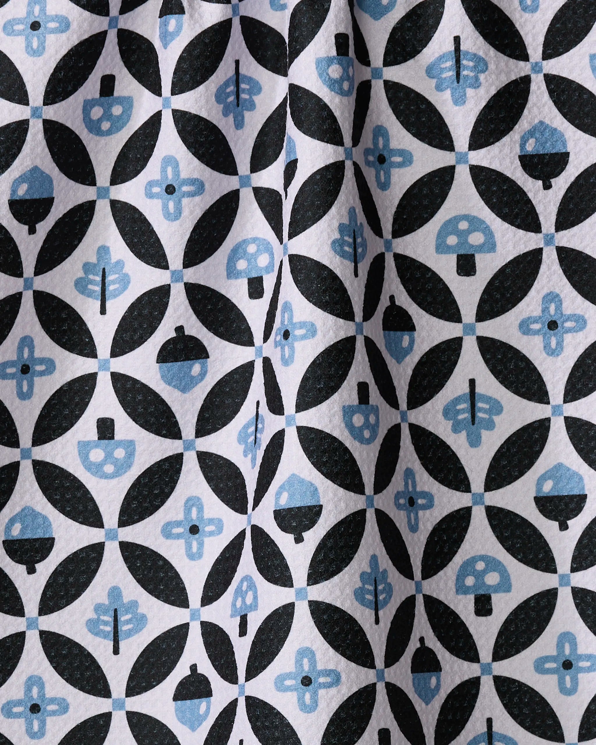 The Forest Findings tea towel by Anette Heiberg features quick-drying fabric with a black and blue geometric print of circles, leaves, acorns, mushrooms, and floral motifs on white. The material has a slightly wrinkled texture.