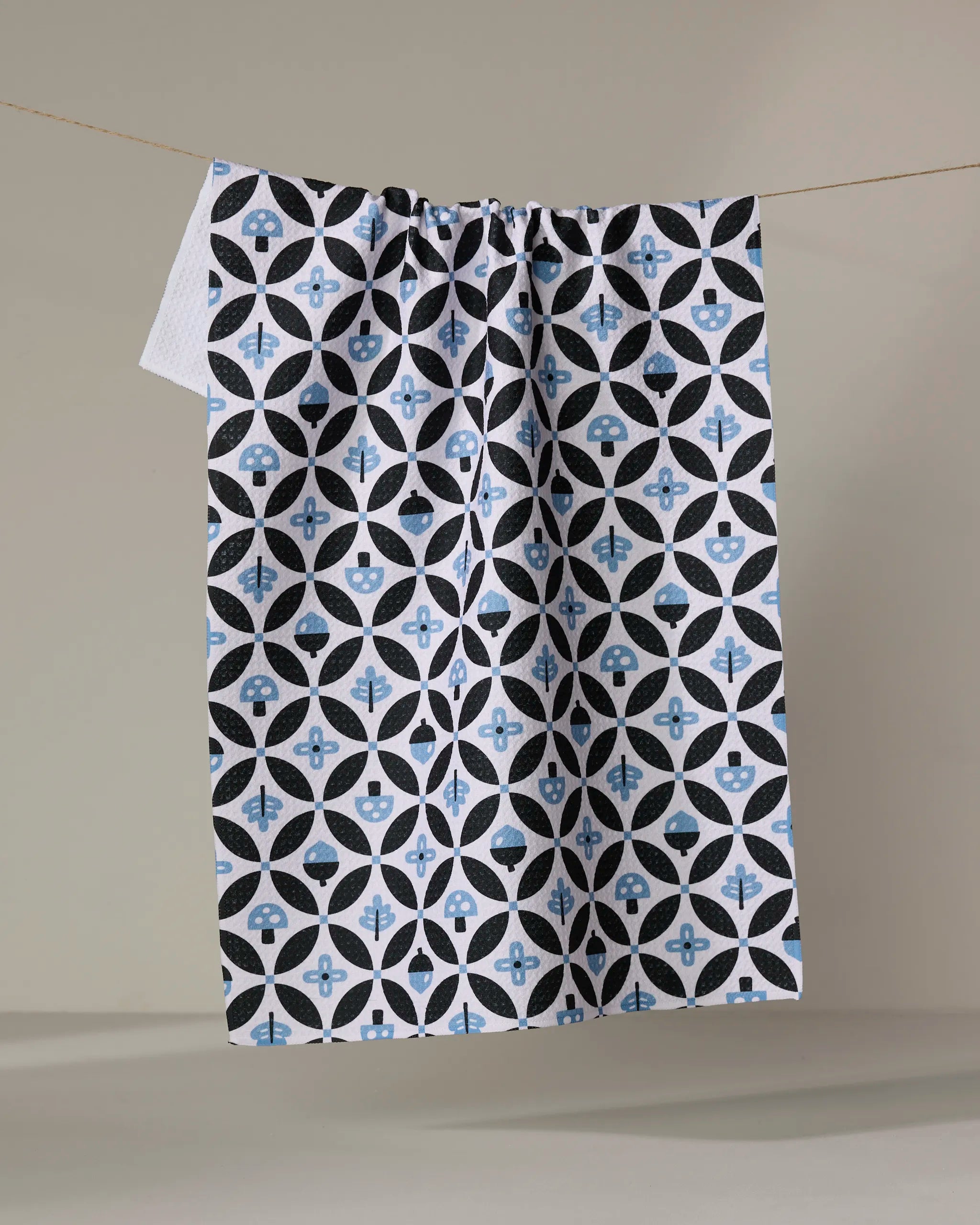 The "Forest Findings" tea towel by Anette Heiberg, featuring a black, blue, and white geometric pattern, hangs drying on a line against a plain beige background.