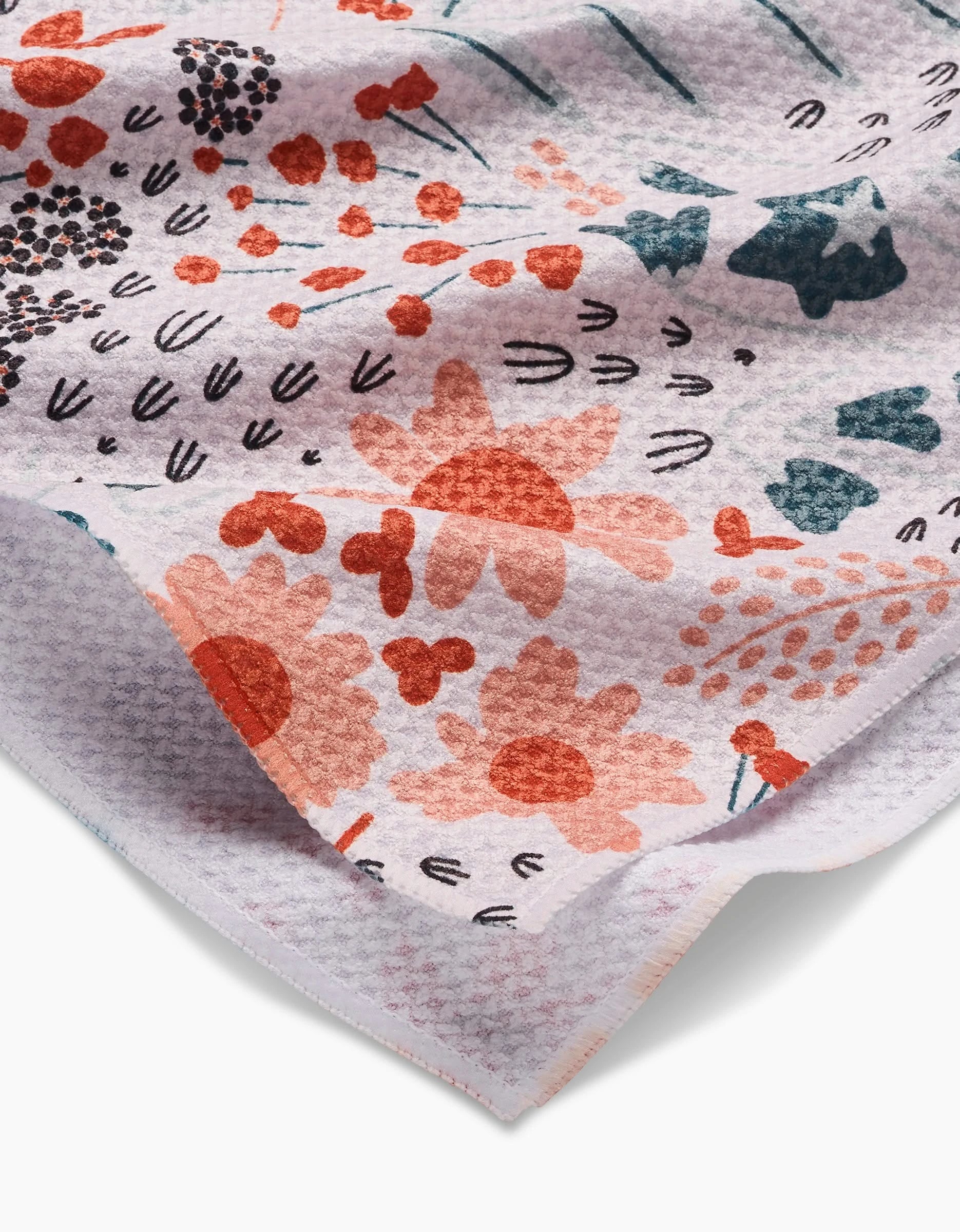 A close-up of Coit Creative’s Forest Foliage tea towel made with Geoweave fabric, showing a colorful floral pattern in coral, orange, teal, and black on a light background. The textured surface and slight fold highlight its unique design.
