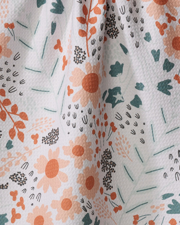 A close-up of Coit Creative's Forest Foliage fabric, featuring a floral pattern in orange, green, and black on white. The recycled material shows flowers, leaves, and abstract shapes with soft folds from gentle gathering.
