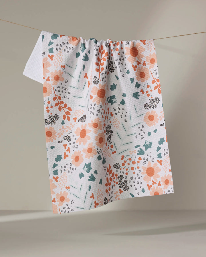 The "Forest Foliage" kitchen towel by Coit Creative, featuring an orange, green, and gray floral and leaf design on white recycled fabric, hangs on a line against a neutral backdrop.