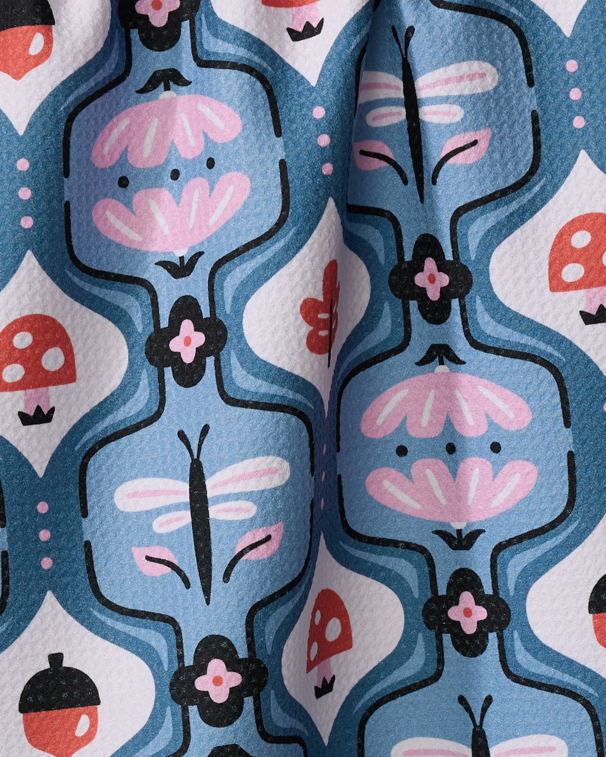 Forest Garden by Anette Heiberg is a colorful recycled kitchen towel with a blue background, featuring black, pink, red, and white dragonflies, mushrooms, acorns, and stylized flowers. The fabric has a slightly wrinkled look.
