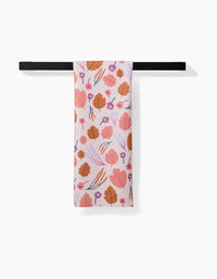 The Forest Leaves pink tea towel by Claire Iglesias, featuring orange and purple floral patterns and crafted from recycled materials, hangs on a black towel bar against a white background.