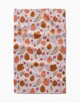 The Forest Leaves tea towel by Claire Iglesias features a Geoweave fabric with brown leaves, small purple flowers, and thin blue and orange lines on a light pink background. There are some slight wrinkles.