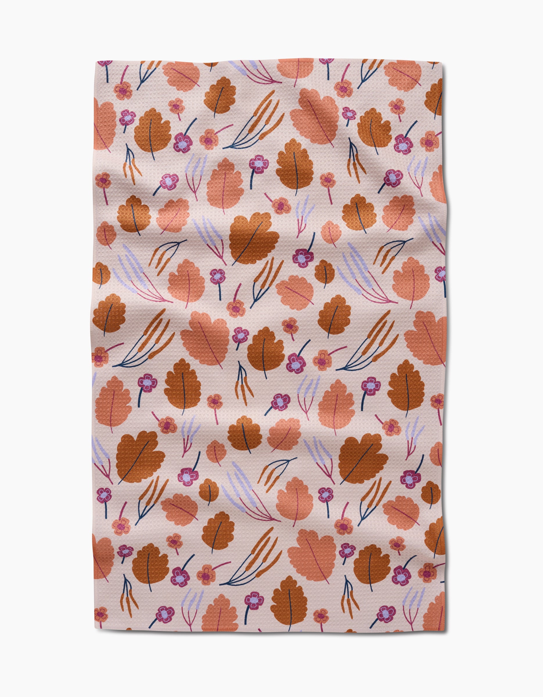 The Forest Leaves tea towel by Claire Iglesias features a Geoweave fabric with brown leaves, small purple flowers, and thin blue and orange lines on a light pink background. There are some slight wrinkles.