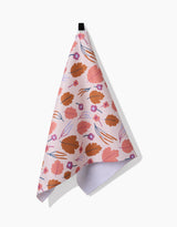 The Forest Leaves tea towel by Claire Iglesias is pink, made from recycled materials, and displays orange leaves with small purple flowers. It hangs from a black loop against a white background.