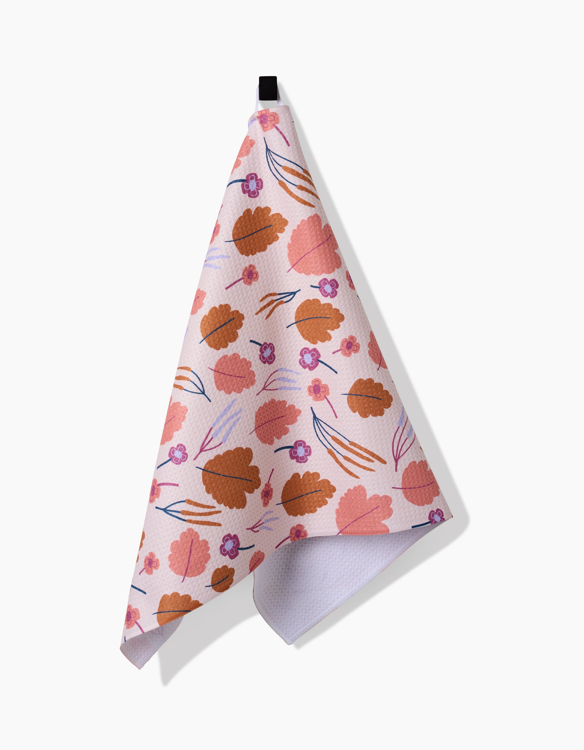 The Forest Leaves tea towel by Claire Iglesias is pink, made from recycled materials, and displays orange leaves with small purple flowers. It hangs from a black loop against a white background.