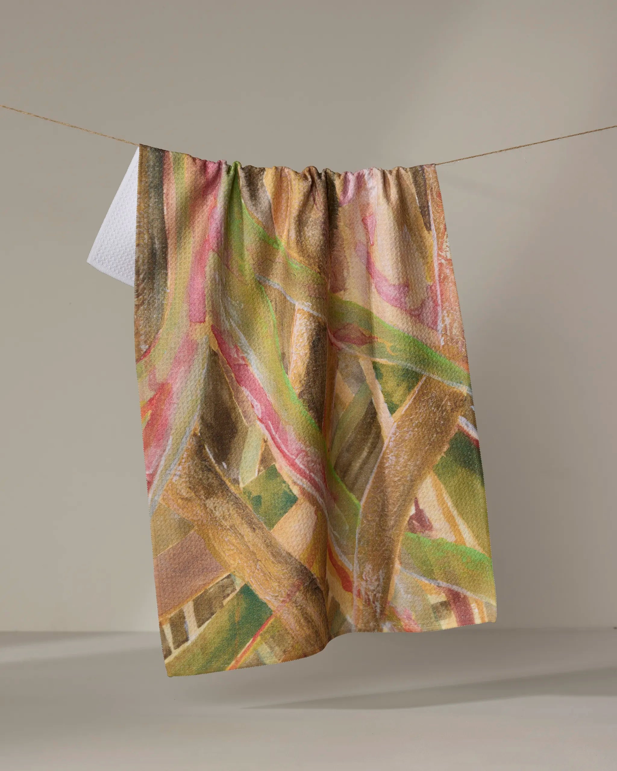 The Forest of Trees tea towel by Taryn Brennan, featuring a colorful abstract pattern in brown, green, pink, and cream, hangs on a clothesline against a plain light background.