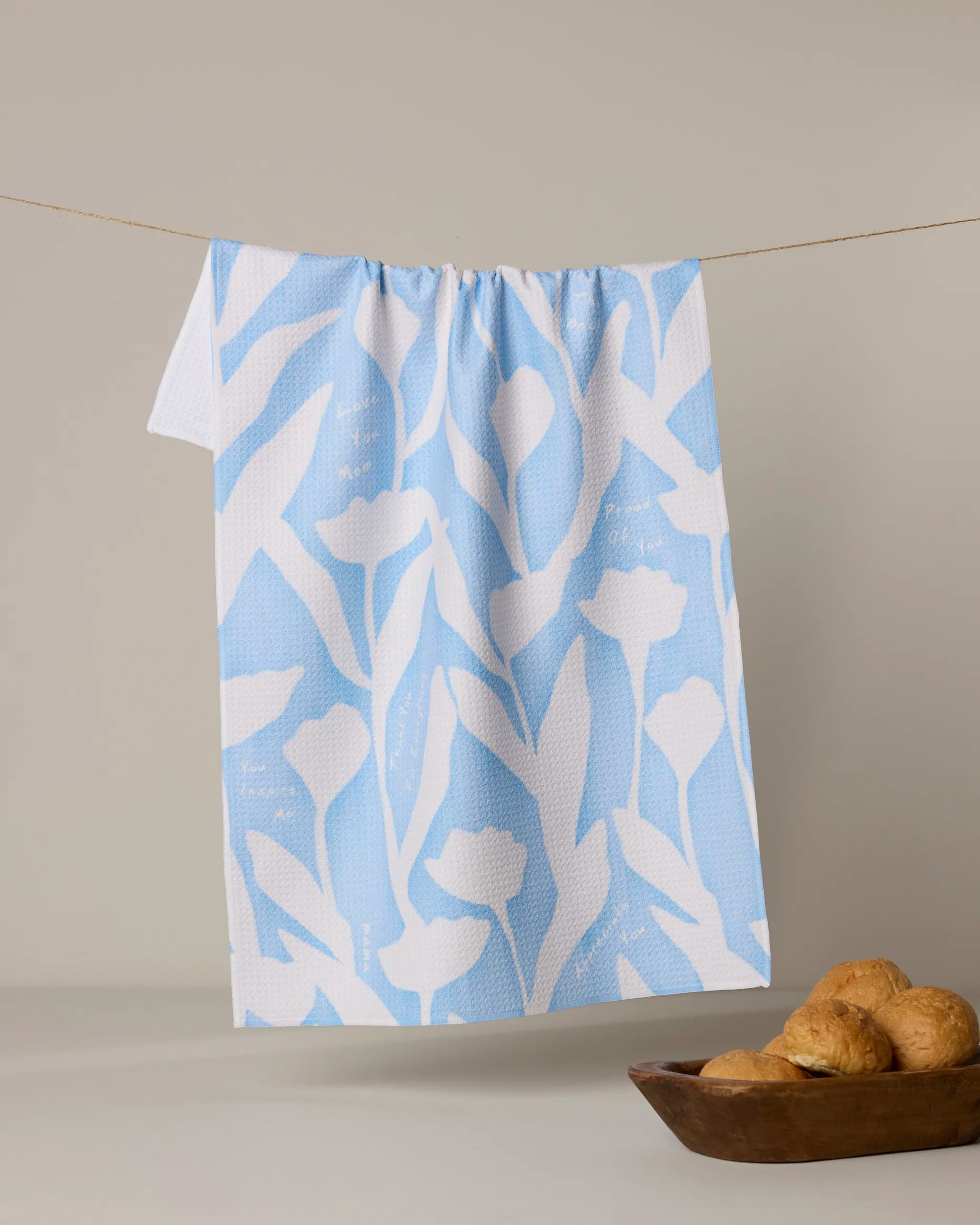 The Forever Grateful tea towel by Geometry features a blue and white floral pattern. Hanging on a clothesline against a light background, it brings charm to any kitchen—especially paired with croissants in a wooden bowl beneath.