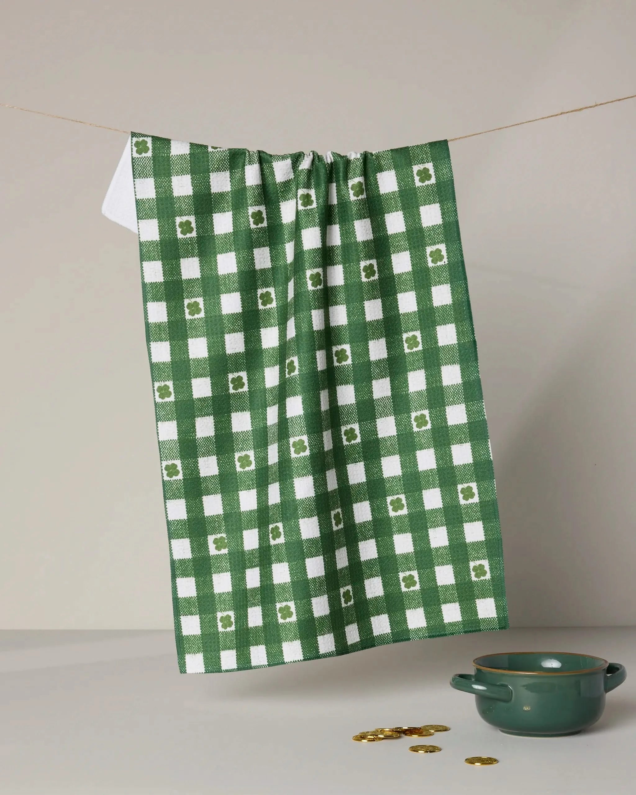 The Fortunate Gingham tea towel by Geometry, featuring a green and white checkered pattern with florals and made from recycled materials, hangs above a green handled bowl on a light surface scattered with gold coins.