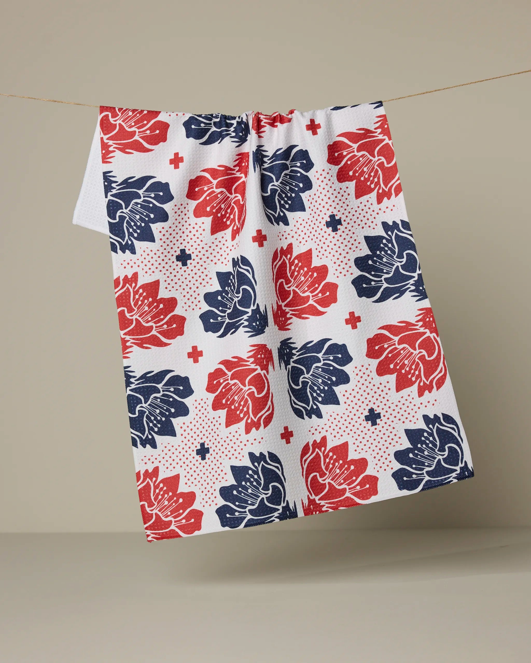 The Founding Floral tea towel by Geometry, featuring a red and blue floral pattern with small red crosses, hangs on a thin line against a plain beige background.
