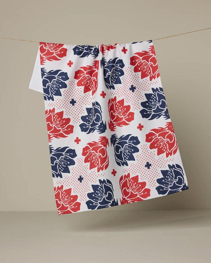 The Founding Floral tea towel by Geometry, featuring a red and blue floral pattern with small red crosses, hangs on a thin line against a plain beige background.