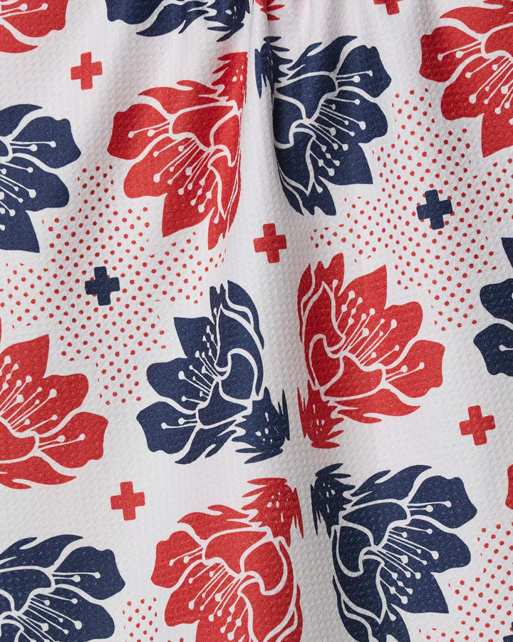 The Geometry Founding Floral tea towel features a bold, eco-friendly white fabric with large red and navy flowers, plus signs, and dotted accents in a symmetrical pattern. Some fabric folds are visible.