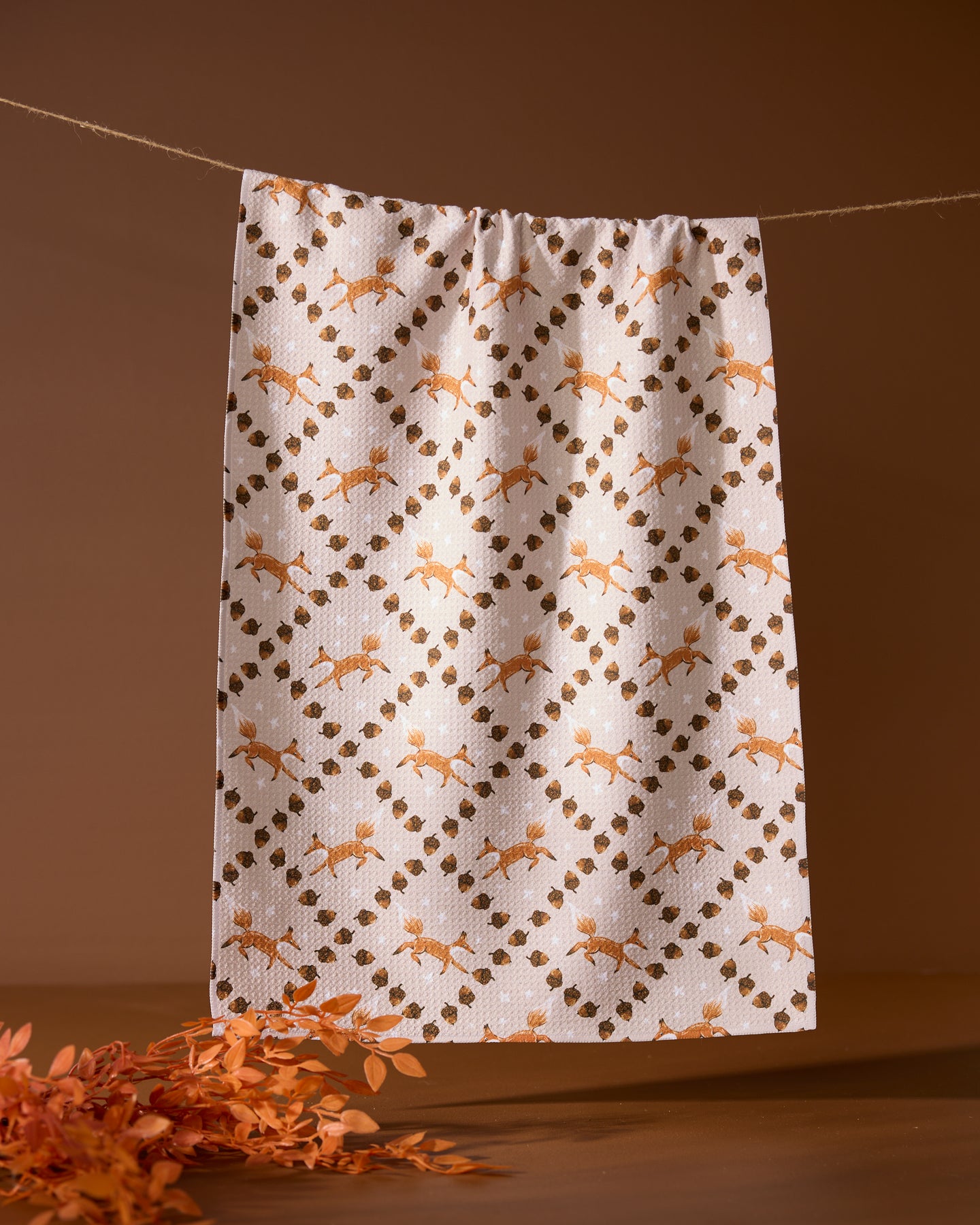 The Geometry Foxes and Twinkles tea towel, featuring an orange and brown floral and leaf design, hangs on a clothesline against a brown background with orange foliage in the bottom left.