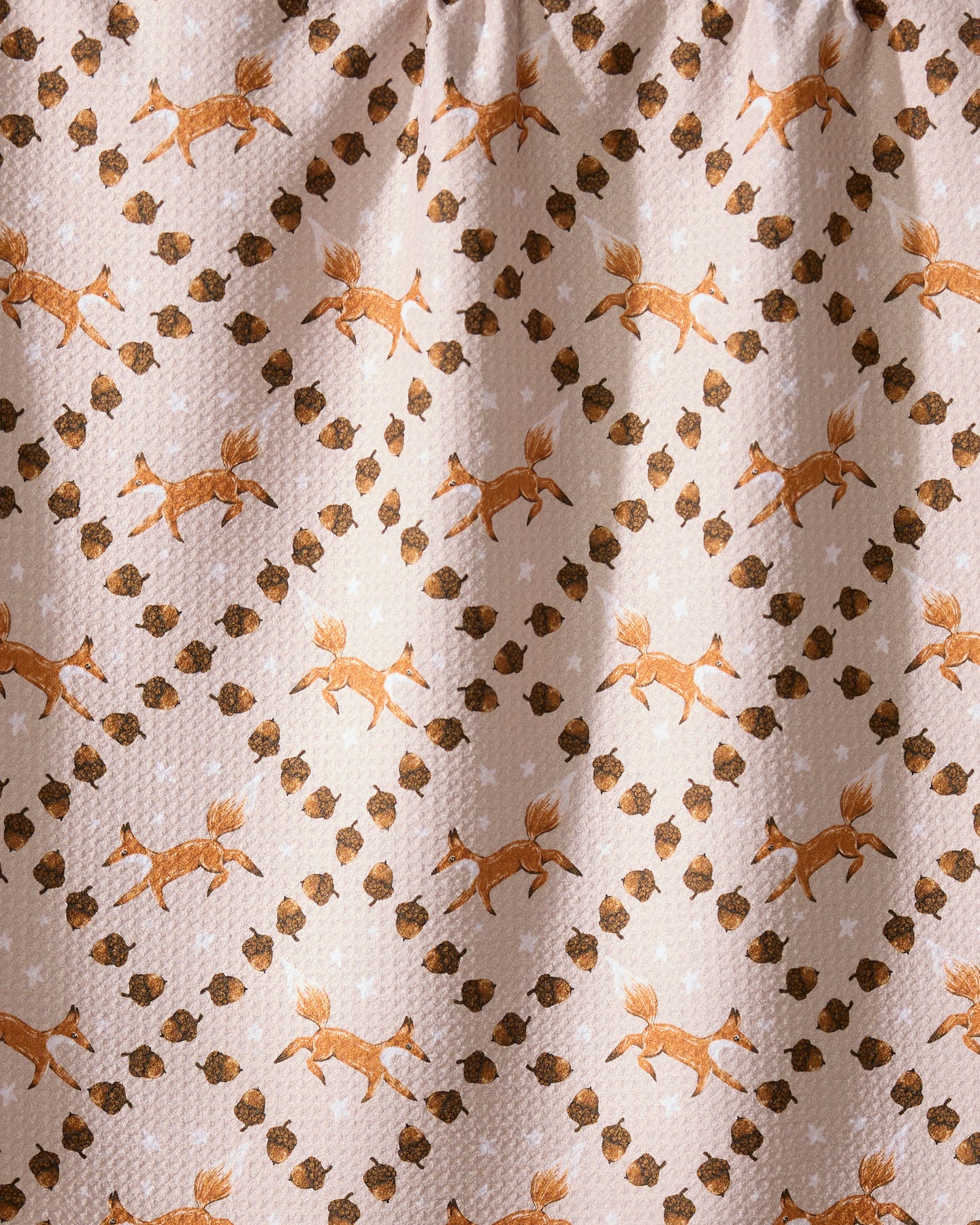 The Foxes and Twinkles tea towel by Geometry features eco-friendly beige fabric with orange foxes, brown acorns, and white stars in a diagonal pattern, plus a subtly textured look and gentle folds at the top.
