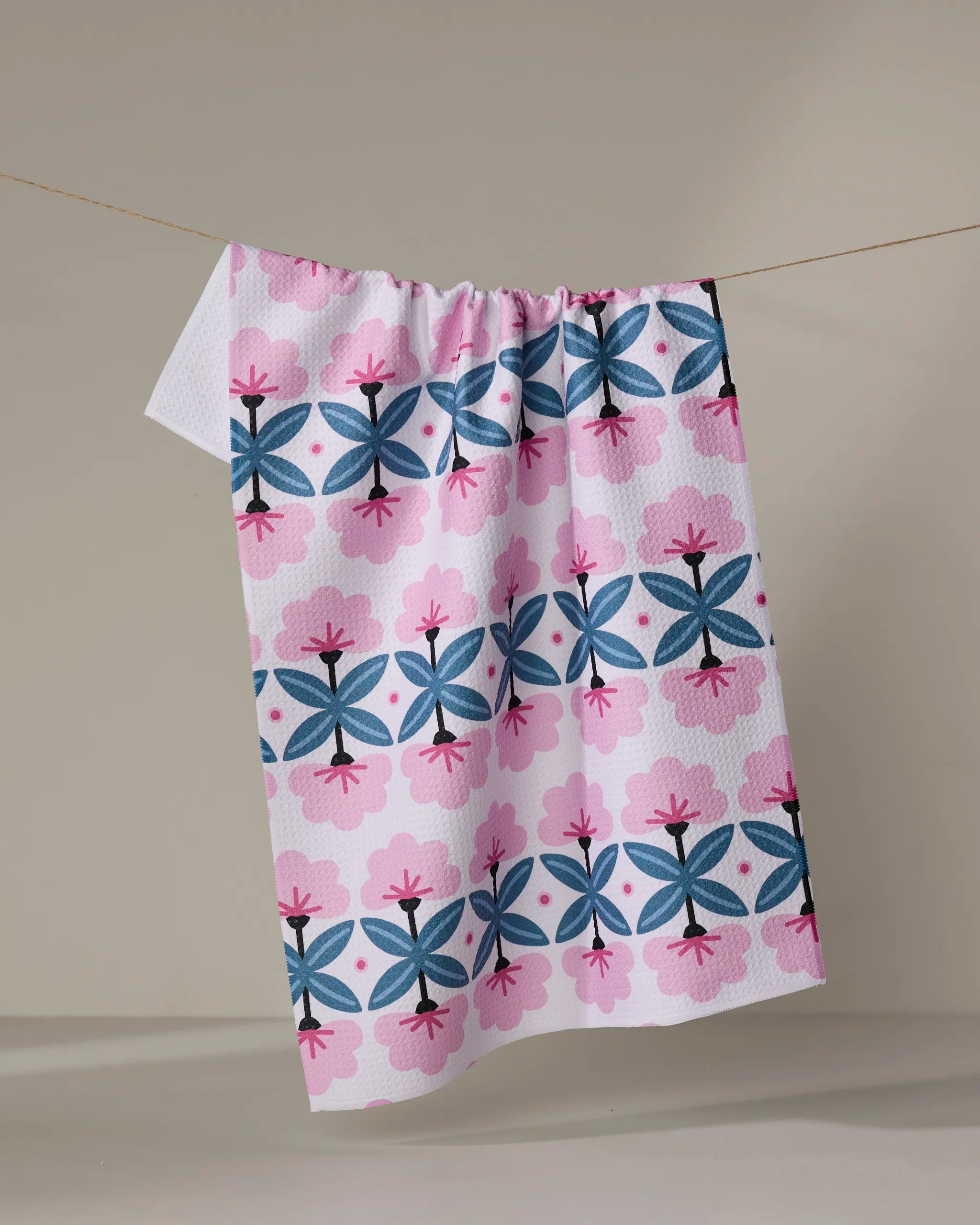 The "Fragrant Flowers" recycled kitchen towel by Anette Heiberg, featuring a pink and blue geometric floral pattern, hangs on a string against a plain light gray background.