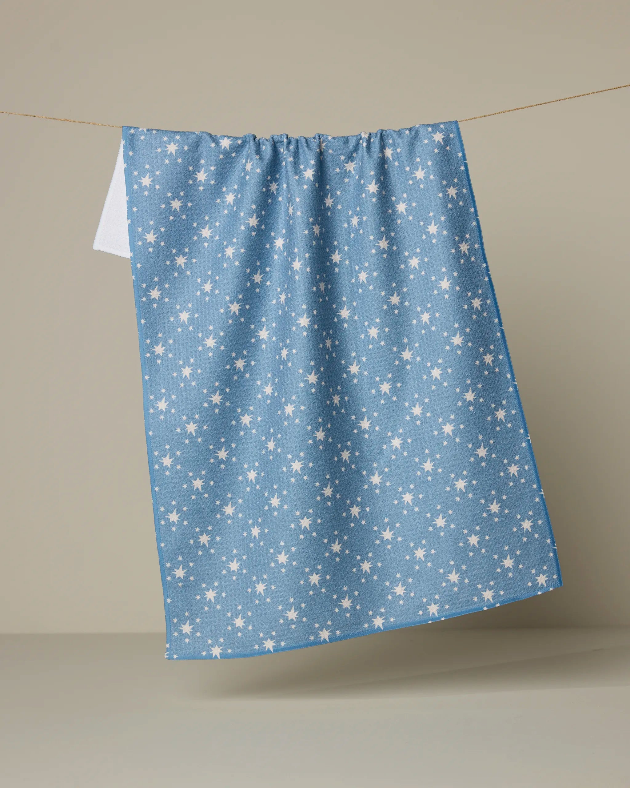 The Geometry Freedom Blue kitchen tea towel, featuring a white star pattern and made from recycled materials, hangs on a clothesline against a plain beige background.