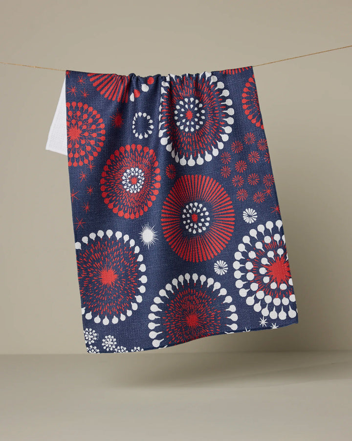 The Geometry Freedom Sparks tea towel in dark blue, featuring red, white, and light blue fireworks circles, hangs on a thin string against a plain beige background.