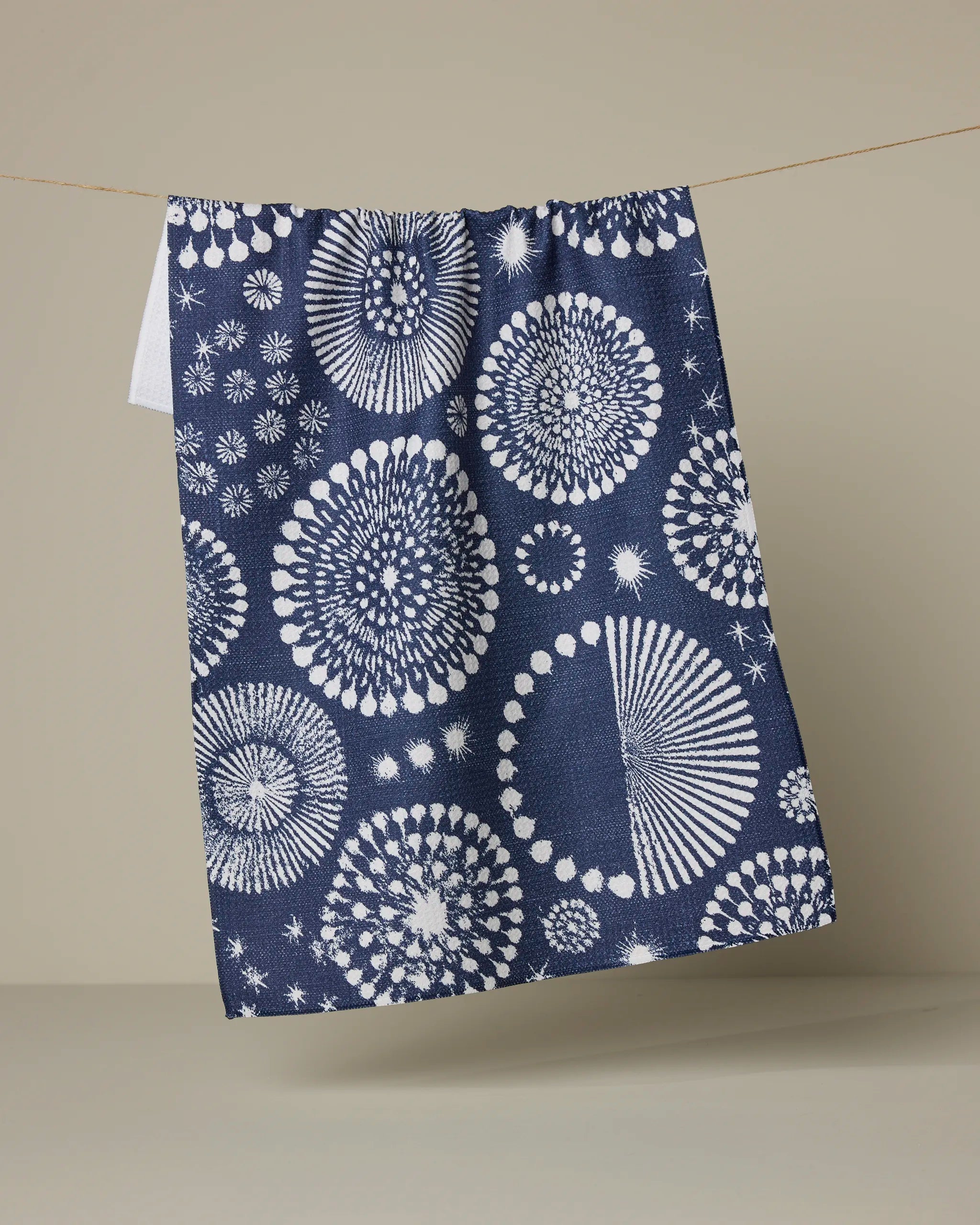The Geometry "Freedom Sparks" tea towel, featuring white circular and starburst designs on navy blue, hangs on a string against a neutral beige background.