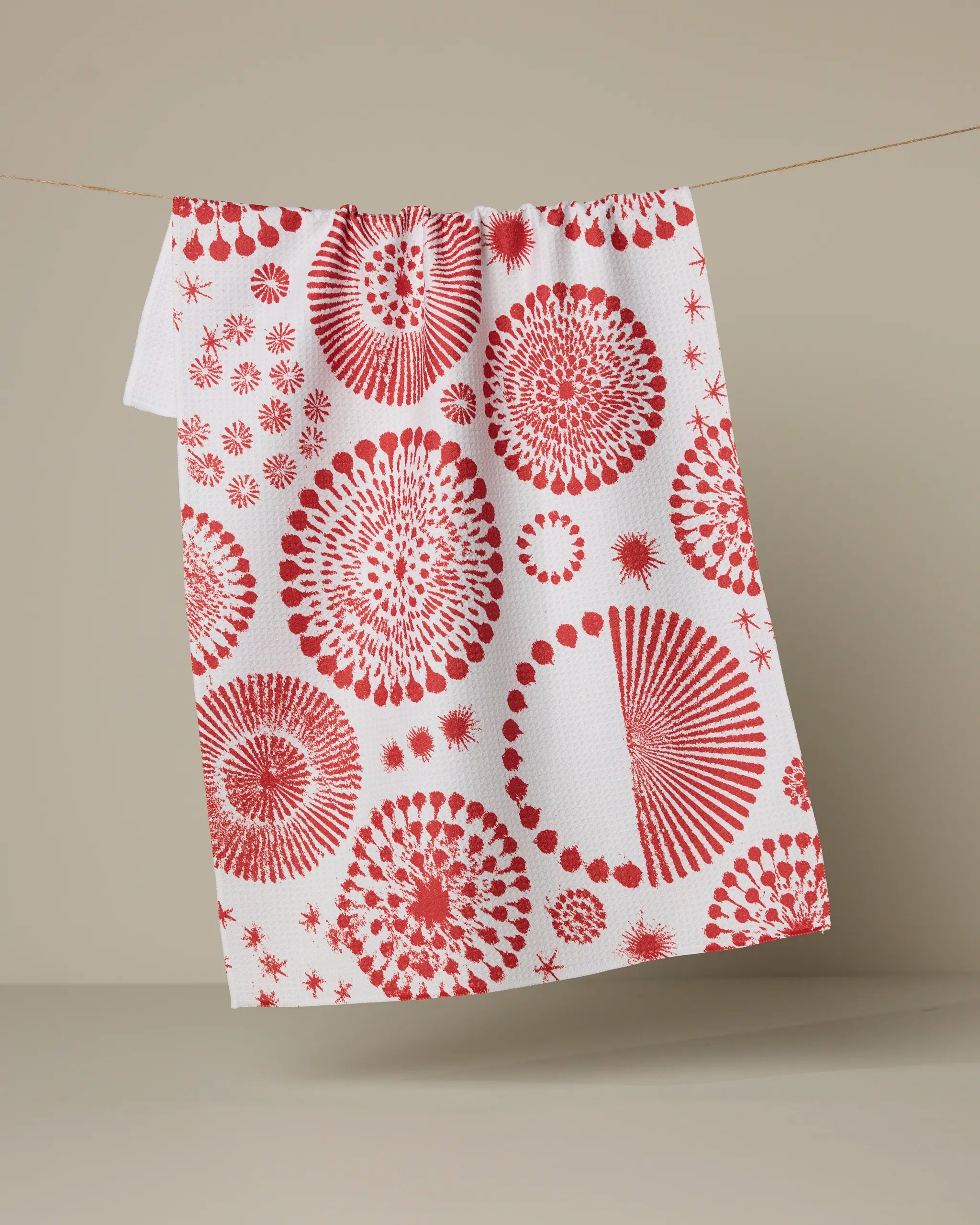 The Geometry "Freedom Sparks" recycled kitchen towel, featuring red geometric and circular patterns on white, hangs on a clothesline against a neutral background.