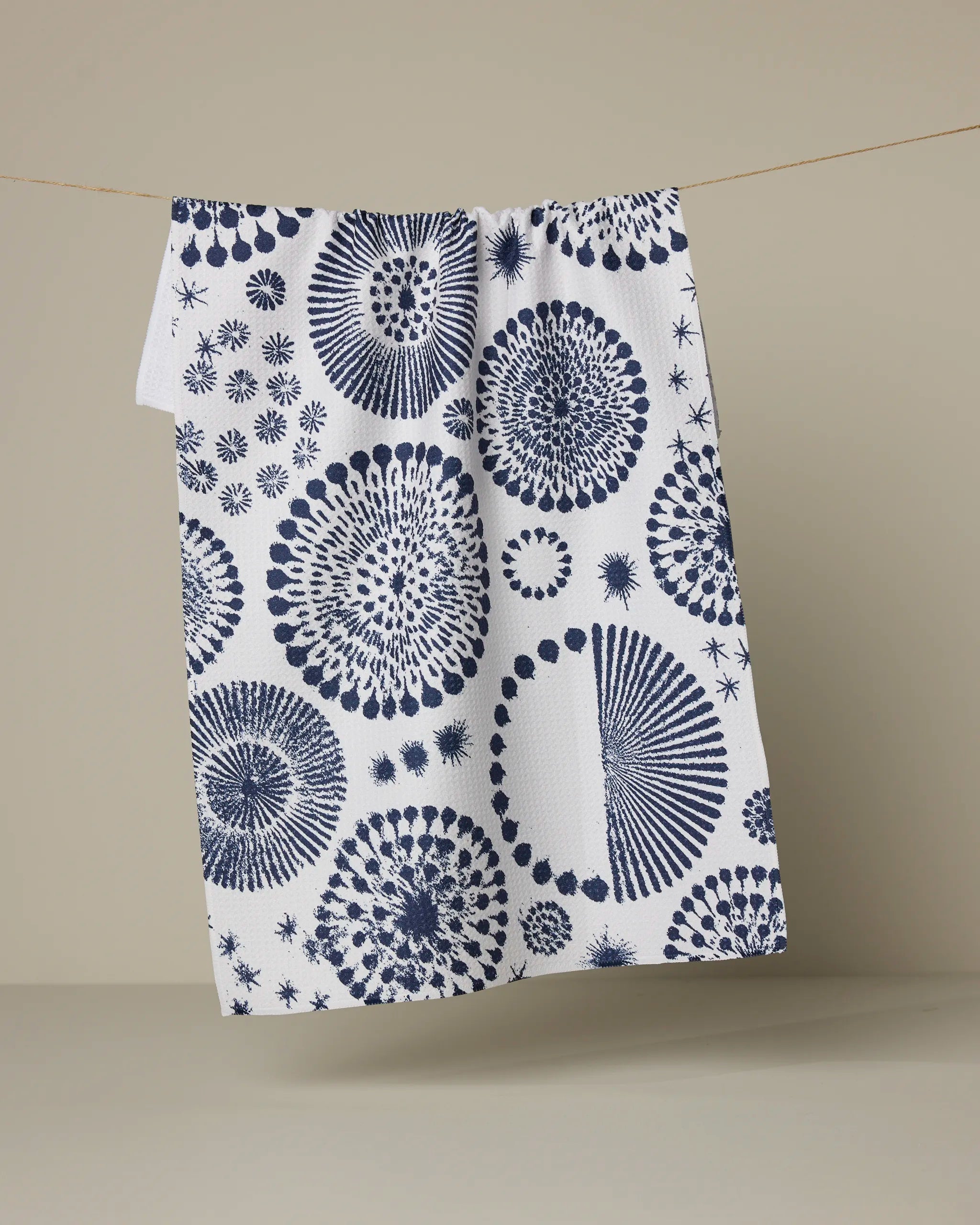The Freedom Sparks recycled kitchen towel by Geometry, featuring blue circular and spiral patterns, hangs on a thin line against a neutral beige background.