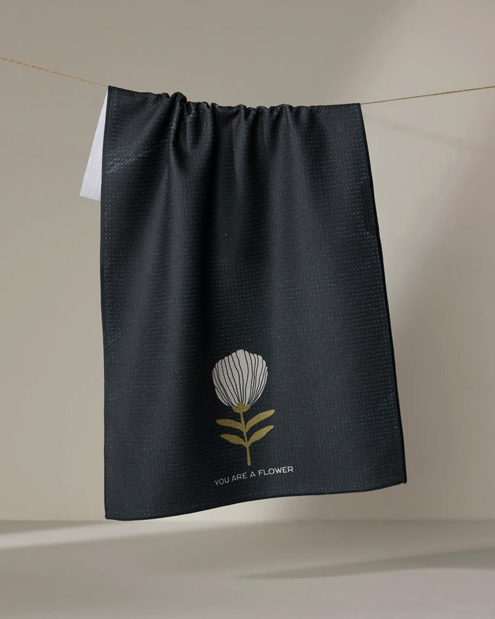 The Freja tea towel by Anna Christian is made from recycled materials, featuring a simple white and yellow flower illustration with the text "YOU ARE A FLOWER," shown hanging against a neutral background.