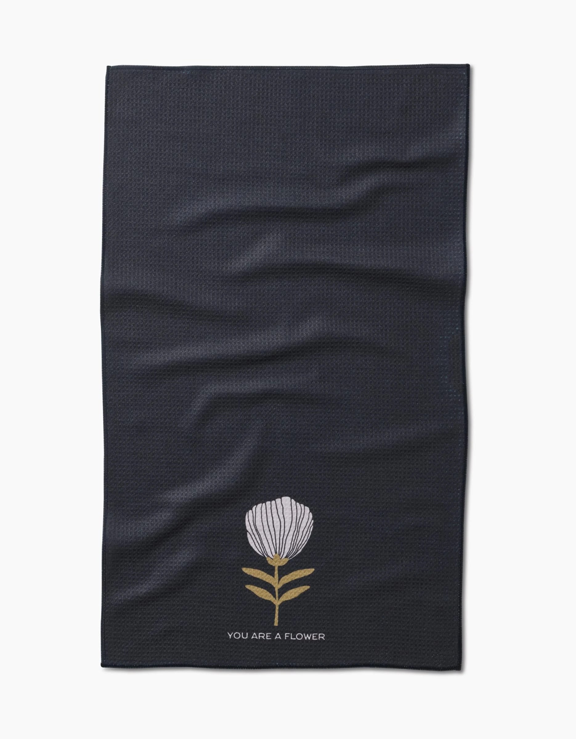 The Freja tea towel by Anna Christian is made from dark Geoweave fabric, featuring a single white flower with green leaves and YOU ARE A FLOWER printed below the illustration.